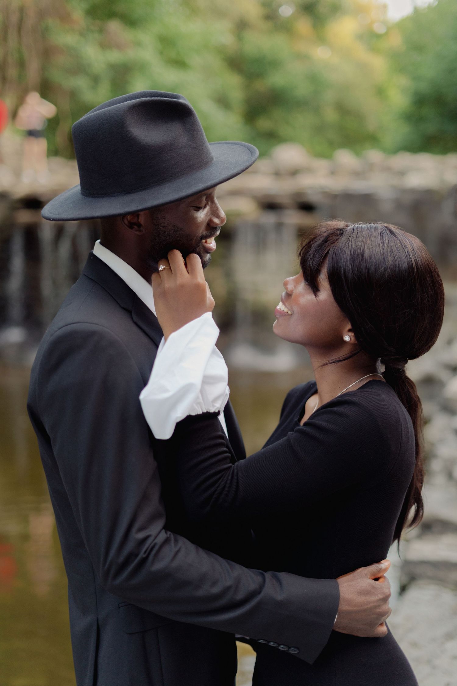 A romantic couple embraces by a pond, with one wearing a black hat and suit while the other wears black attire.