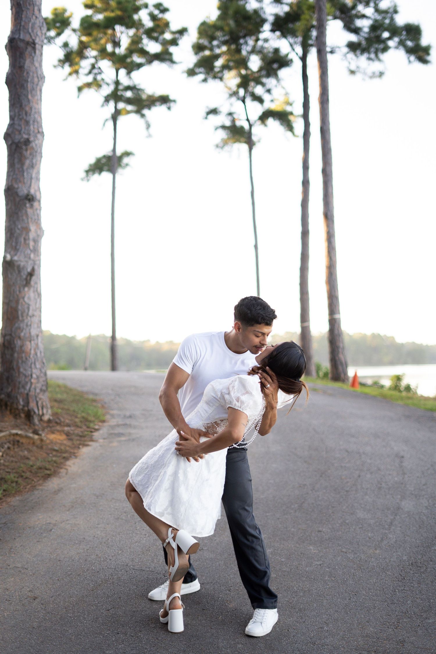 Couple in white outfits dances playfully on path lined with tall pine trees.