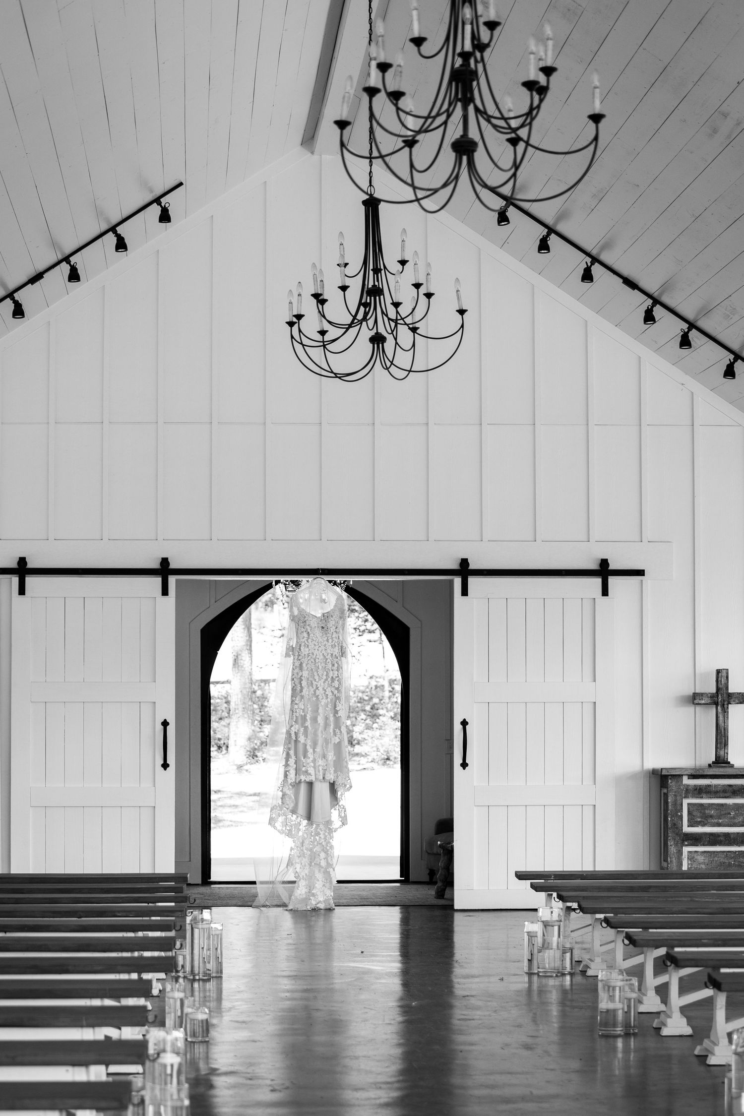 Interior of a modern white chapel with wooden pews and elegant black chandeliers hanging from a vaulted ceiling.