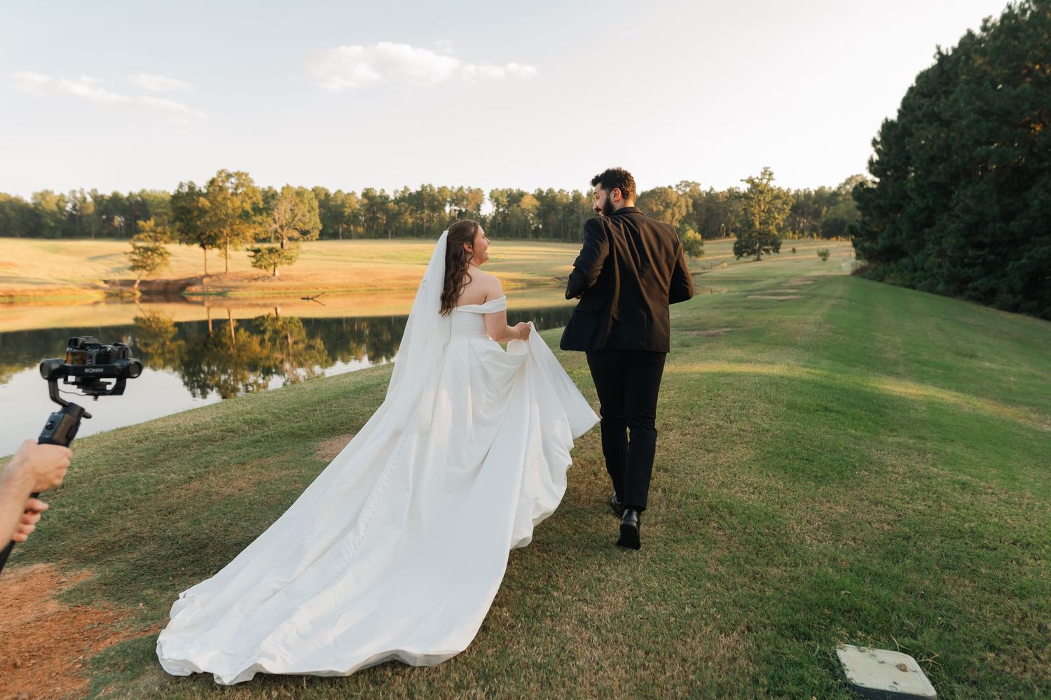 A couple in wedding attire walks across a scenic golf course during golden hour.