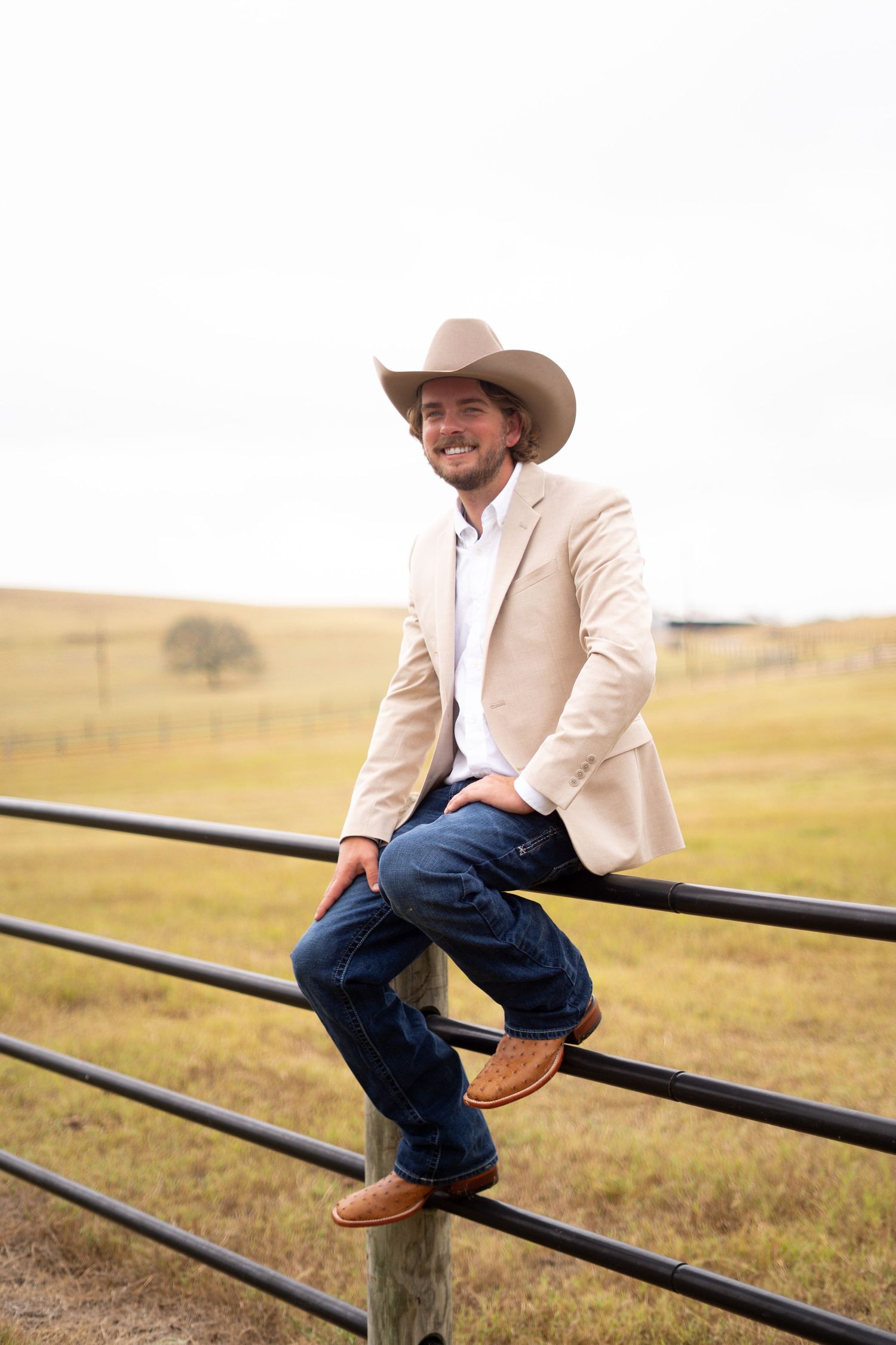 A person in a beige blazer and cowboy hat sits casually on a wooden fence with rolling hills in background.