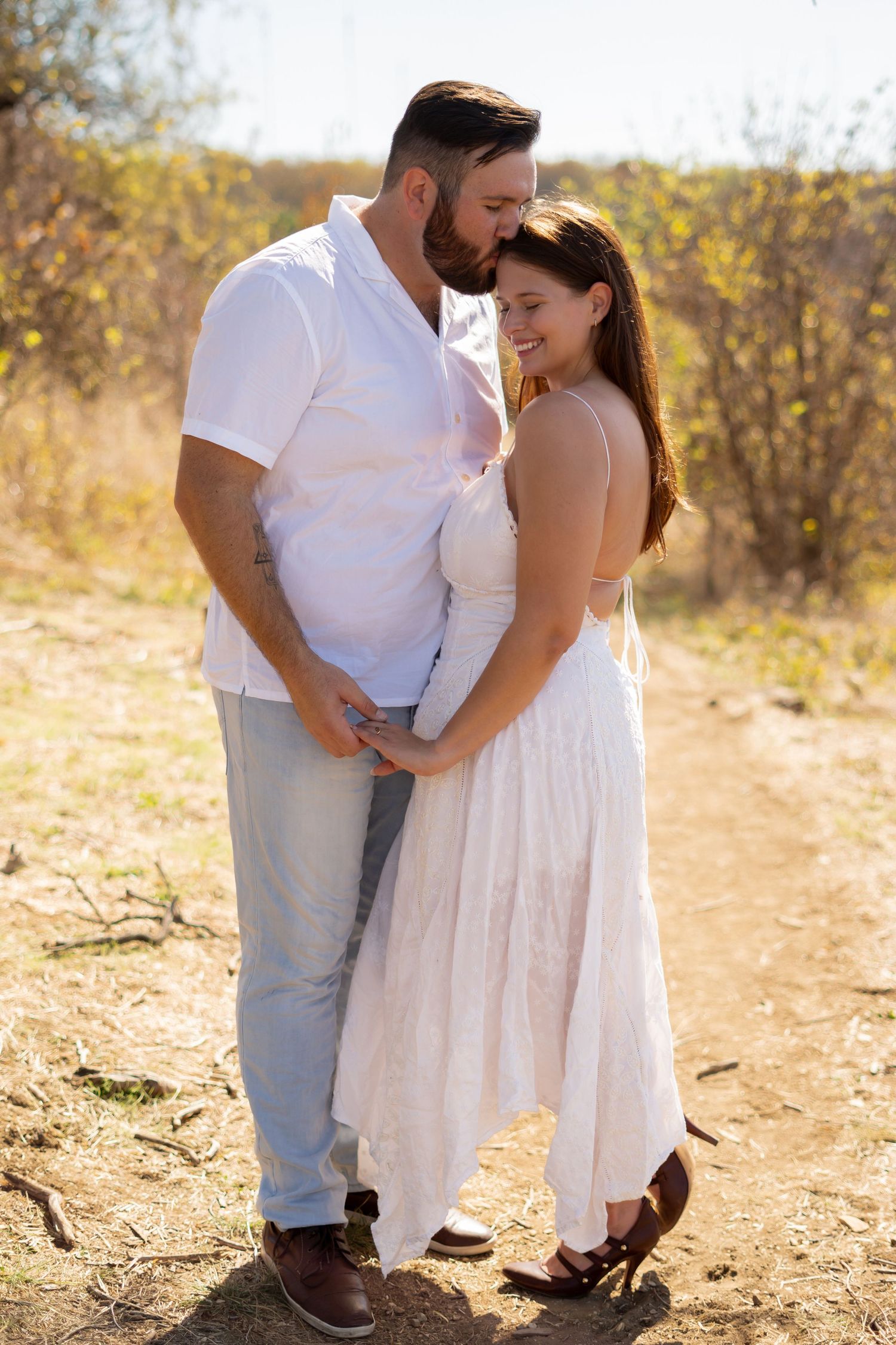 A romantic couple in white outfits embraces during a golden hour photoshoot in a desert landscape.