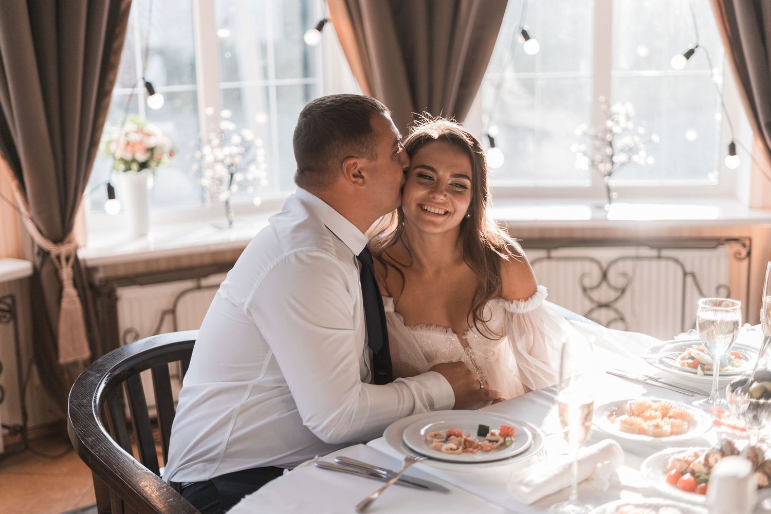 Romantic couple sharing intimate moments at a restaurant table with candles and curtains in the background.