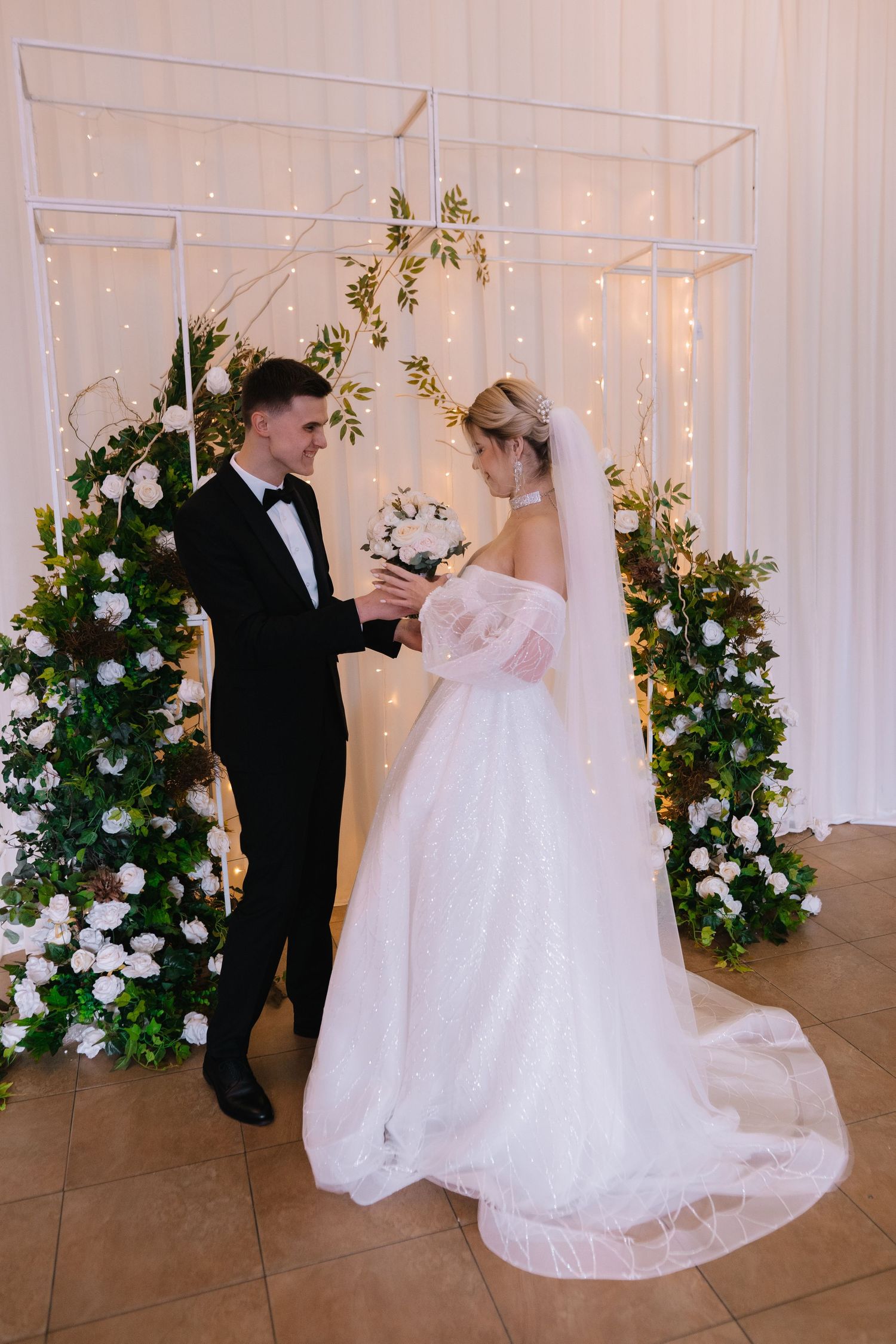 Romantic wedding couple dancing together in an elegant white room decorated with floral arrangements and twinkling lights.