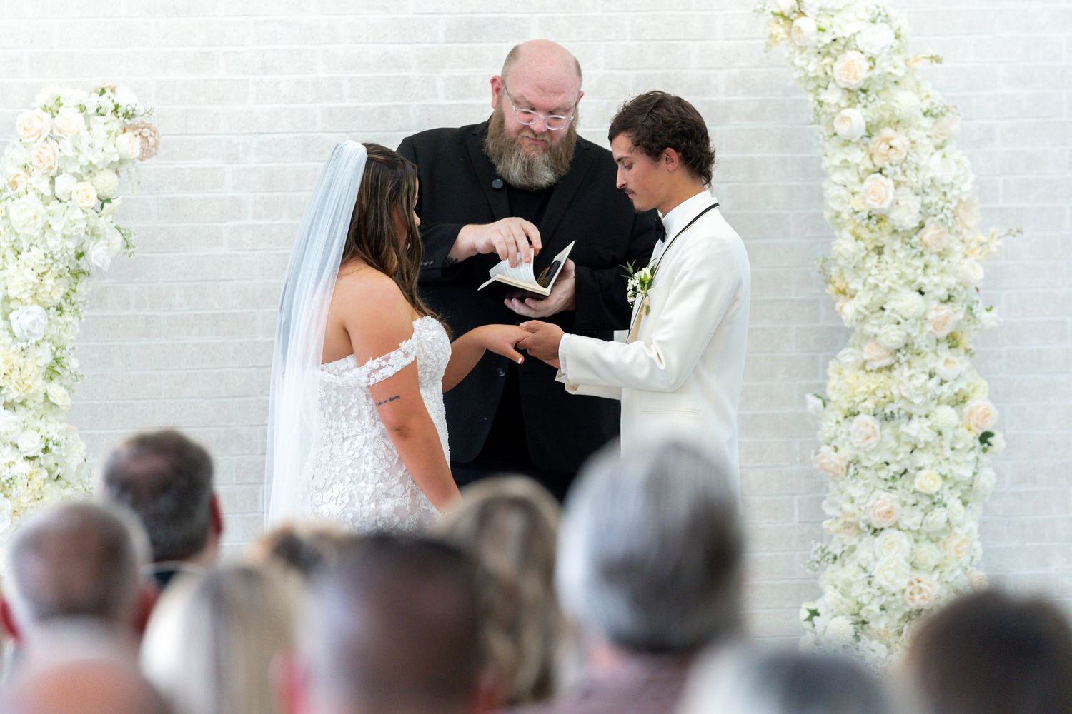 A couple stands at a wedding ceremony altar decorated with white floral arrangements as guests look on.
