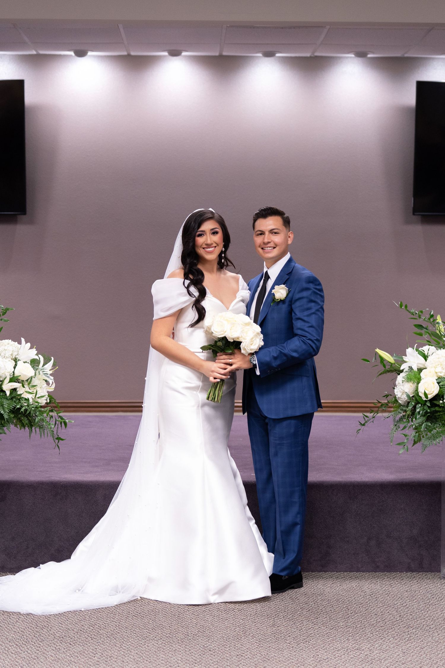 A bride in white dress and groom in navy suit pose together at their wedding ceremony with floral arrangements.