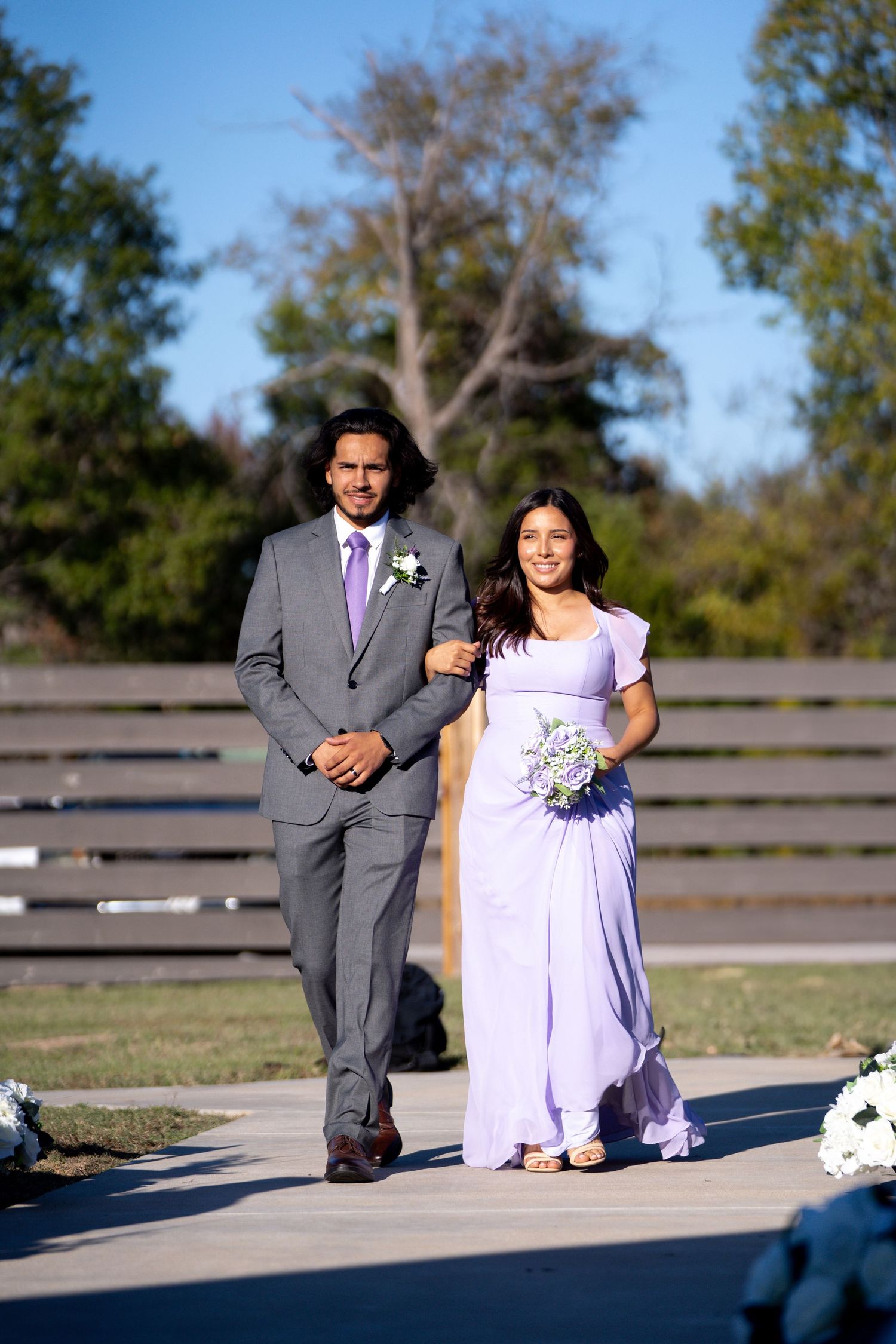 Wedding party member in grey suit escorts bridesmaid in lavender dress down outdoor pathway.