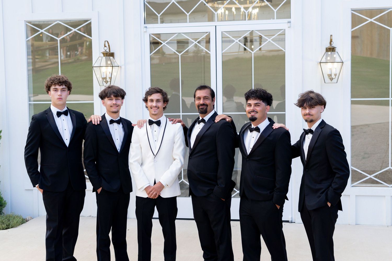 A group of young men in formal black and white tuxedos pose together at an elegant venue with glass doors.