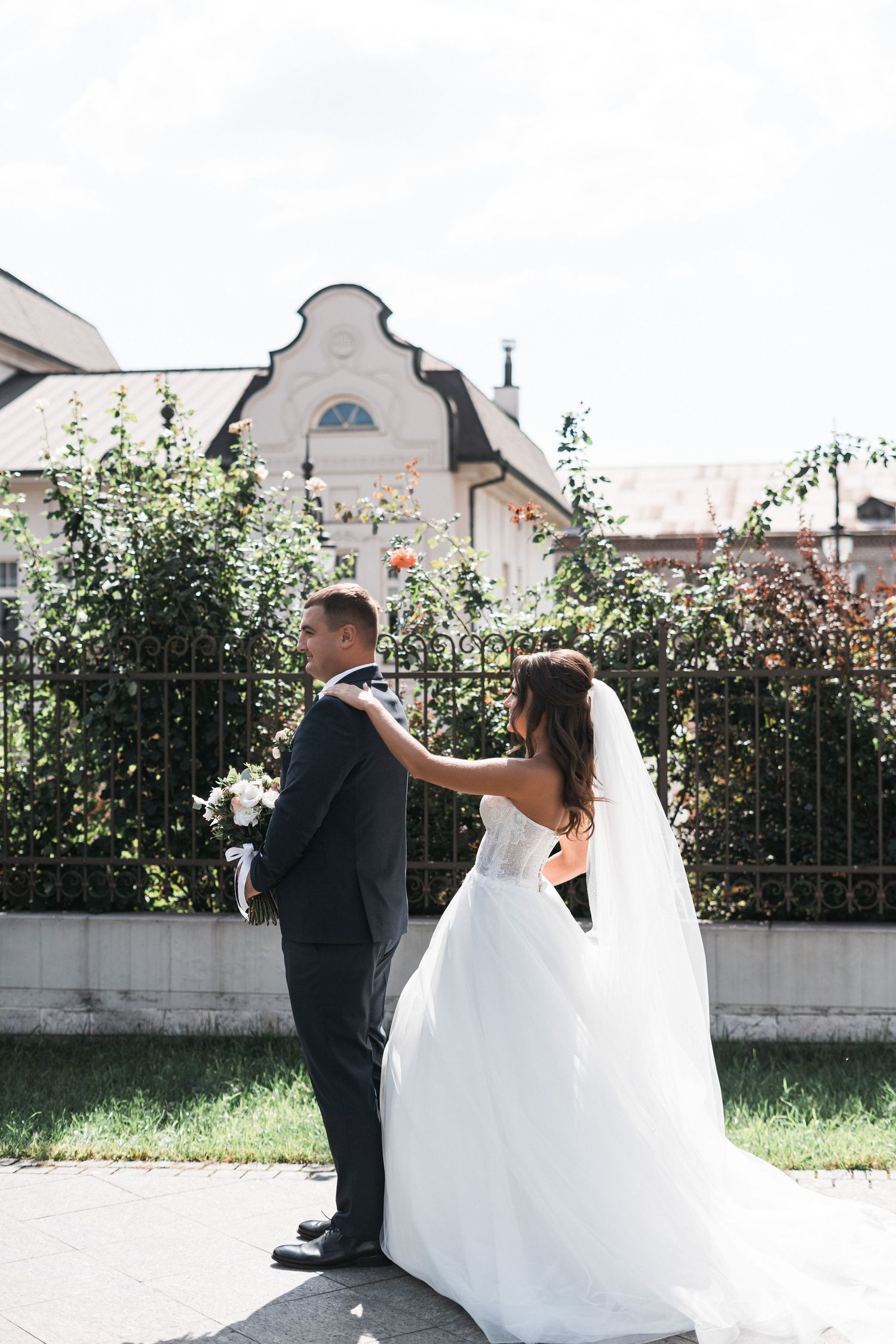 A bride and groom share a first look moment in front of a white building with flowering bushes and an iron fence.