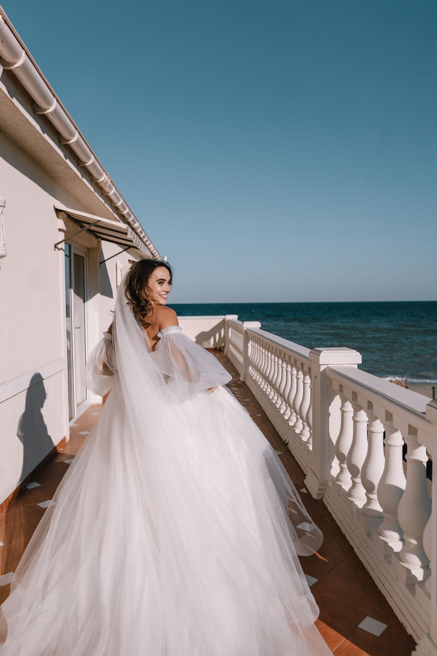 A bride in a flowing white wedding dress poses along a seaside balcony with white railings on a sunny day.
