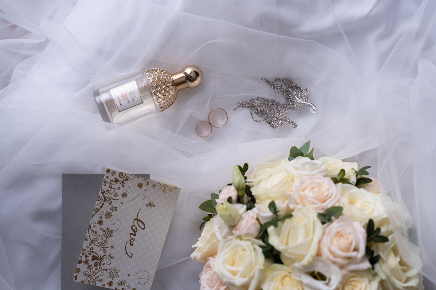 Elegant wedding accessories with white roses, lace veil, and gold jewelry arranged on a soft gray background.