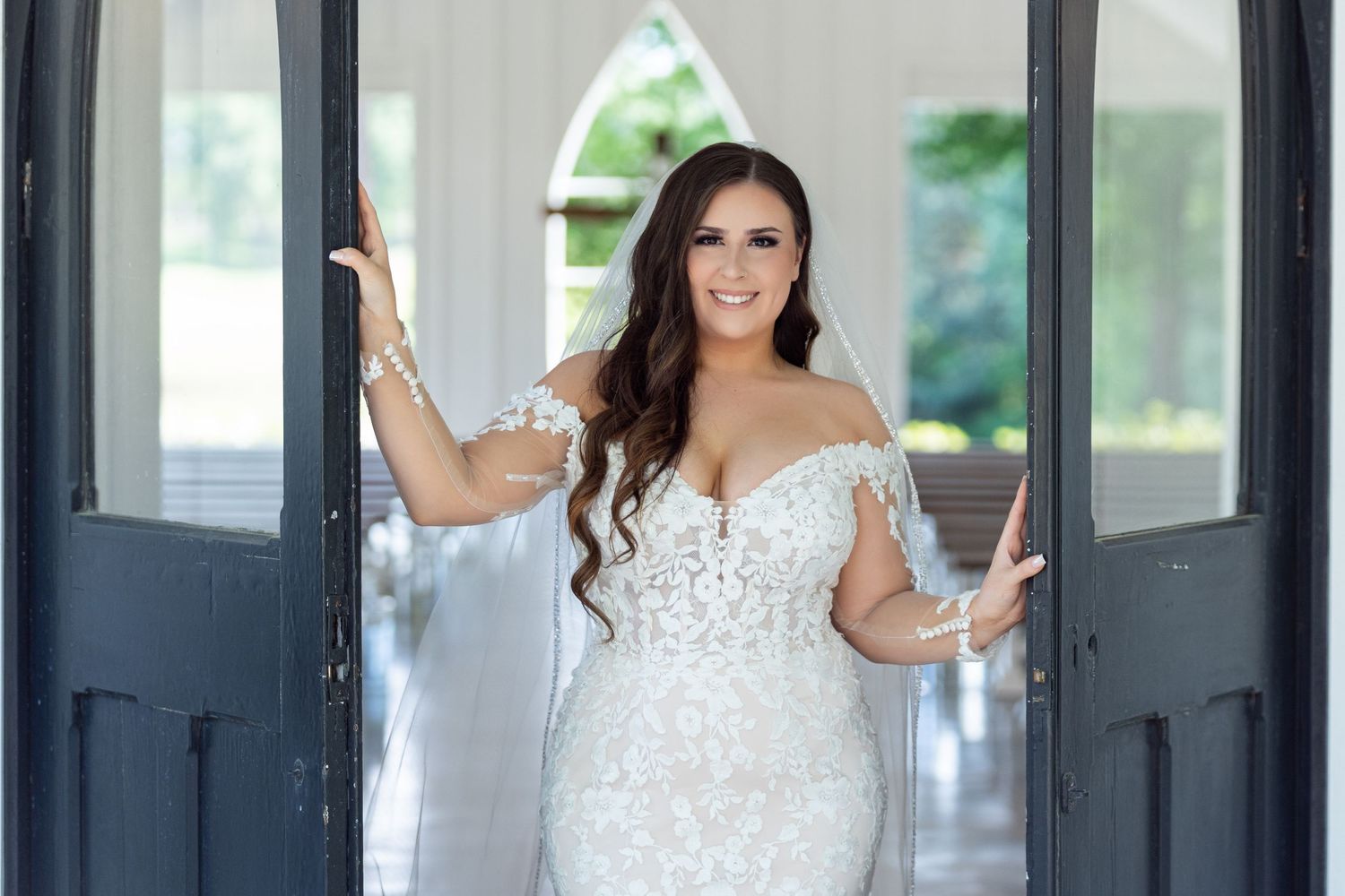A bride in a fitted off-shoulder lace wedding gown stands between elegant dark doors in a bright entryway.