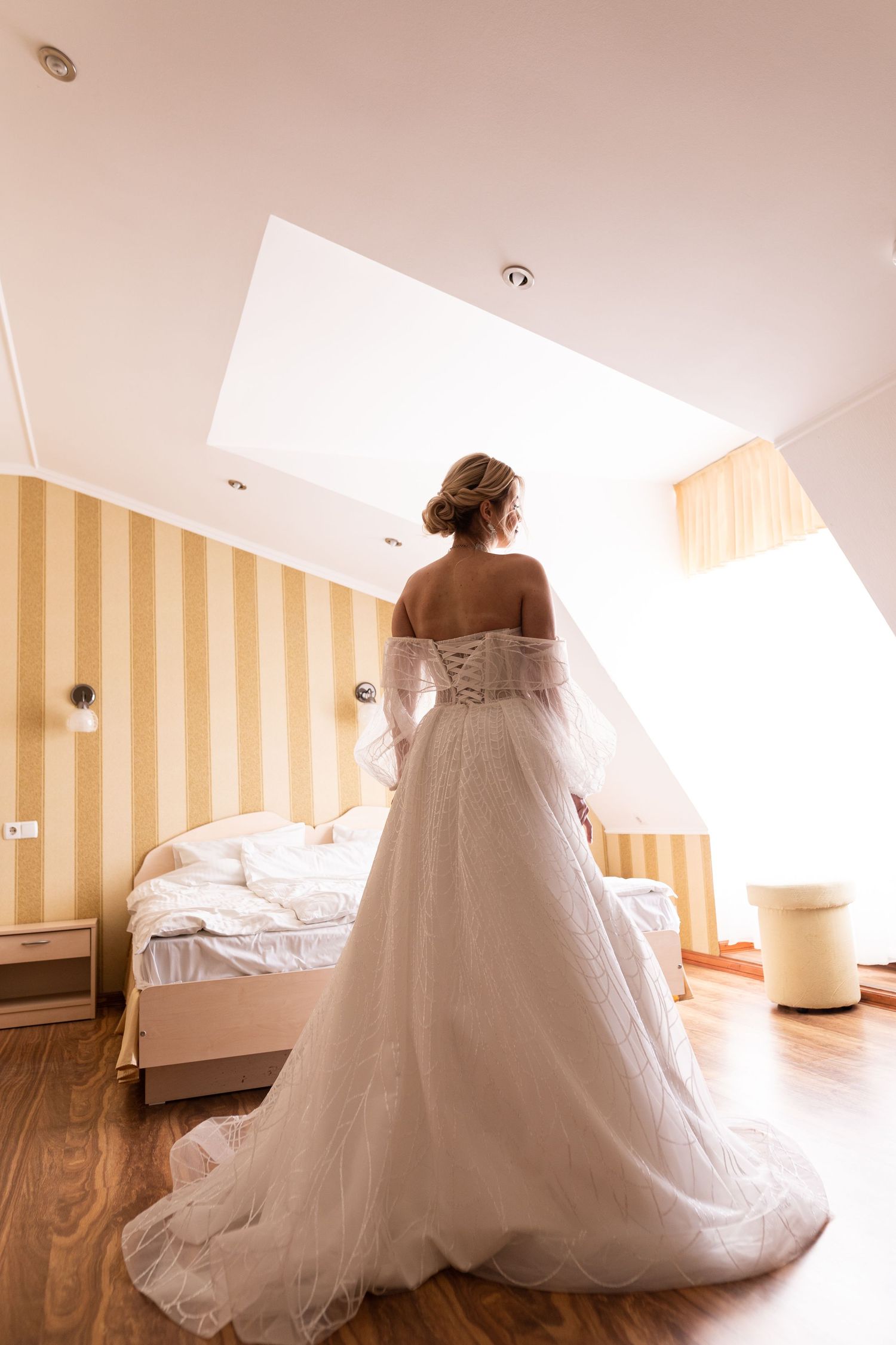 Back view of a wedding dress with corset detail and flowing tulle skirt in a sunlit bedroom with striped walls.