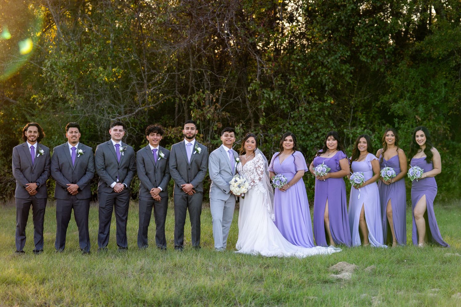 Wedding party in purple and gray attire poses together on grassy field.