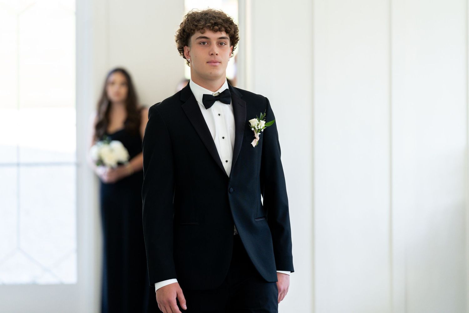 A groom in a classic black tuxedo with bow tie stands in an elegant white room during a formal wedding event.