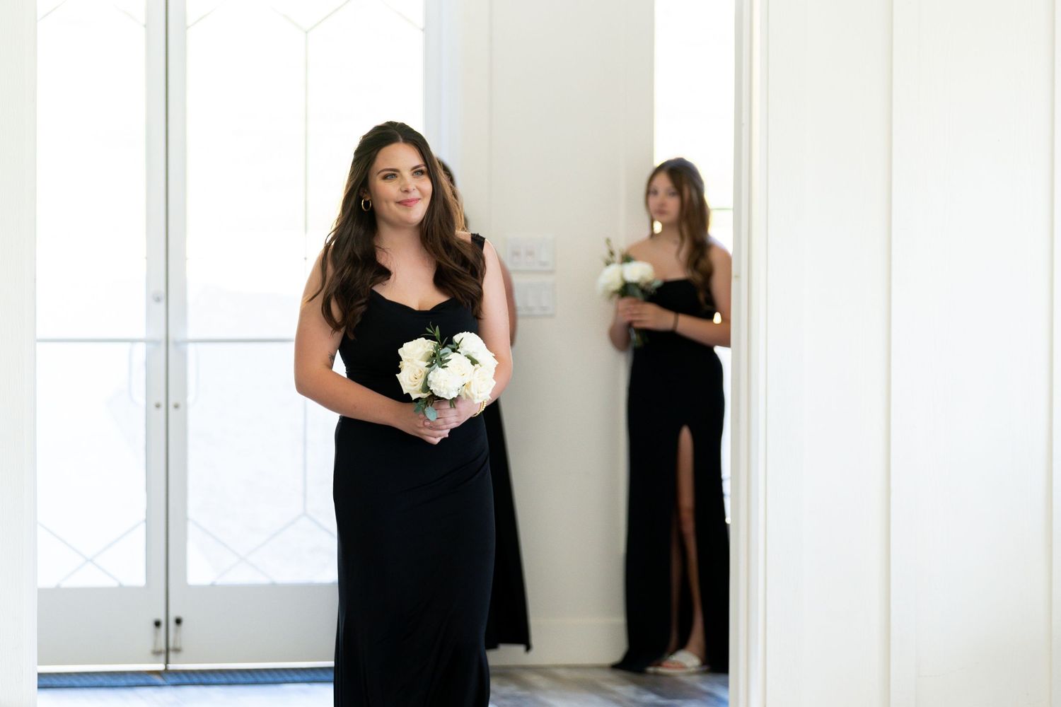 Two people in elegant black formal dresses stand in a bright white room, one holding a white floral bouquet.