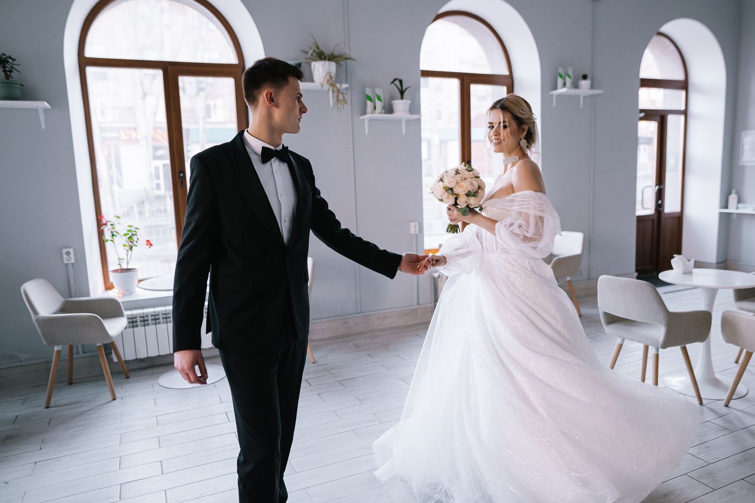 A couple in formal wedding attire dance together in a bright room with arched windows and white decor.