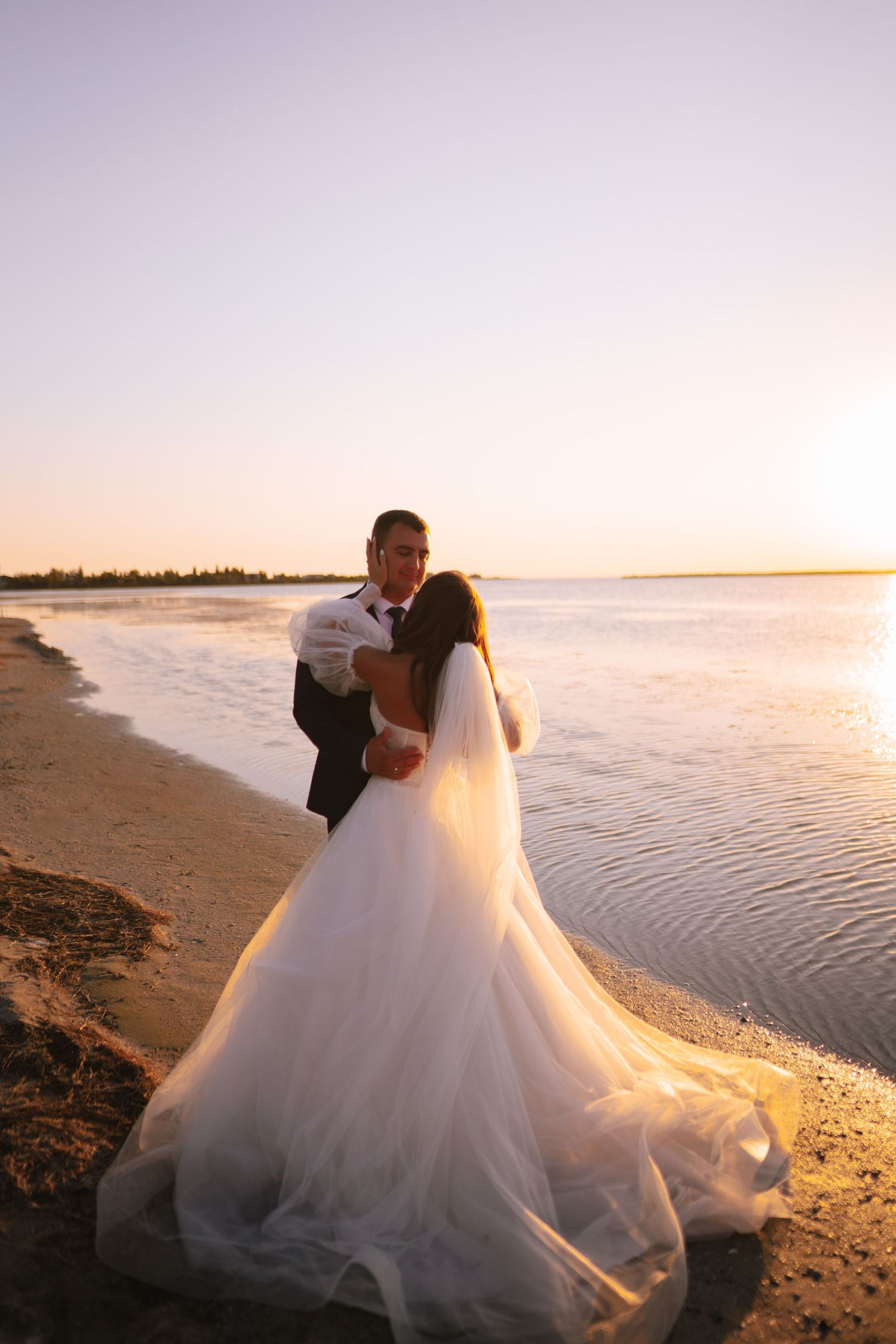 A couple in wedding attire embraces on a sandy beach at sunset with ocean waves in the background.
