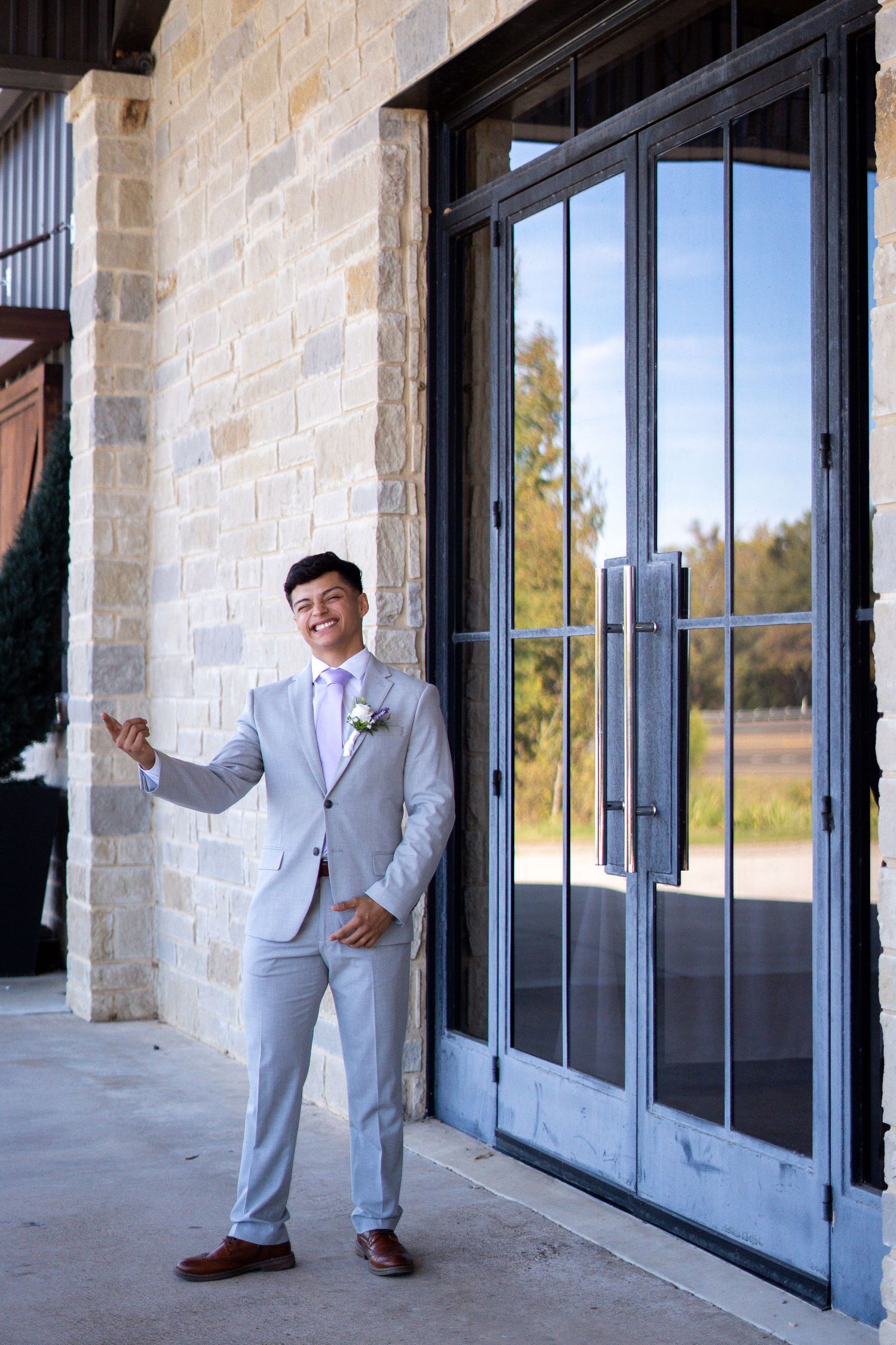 A groom in a light gray suit poses playfully in front of glass windows at a modern venue.