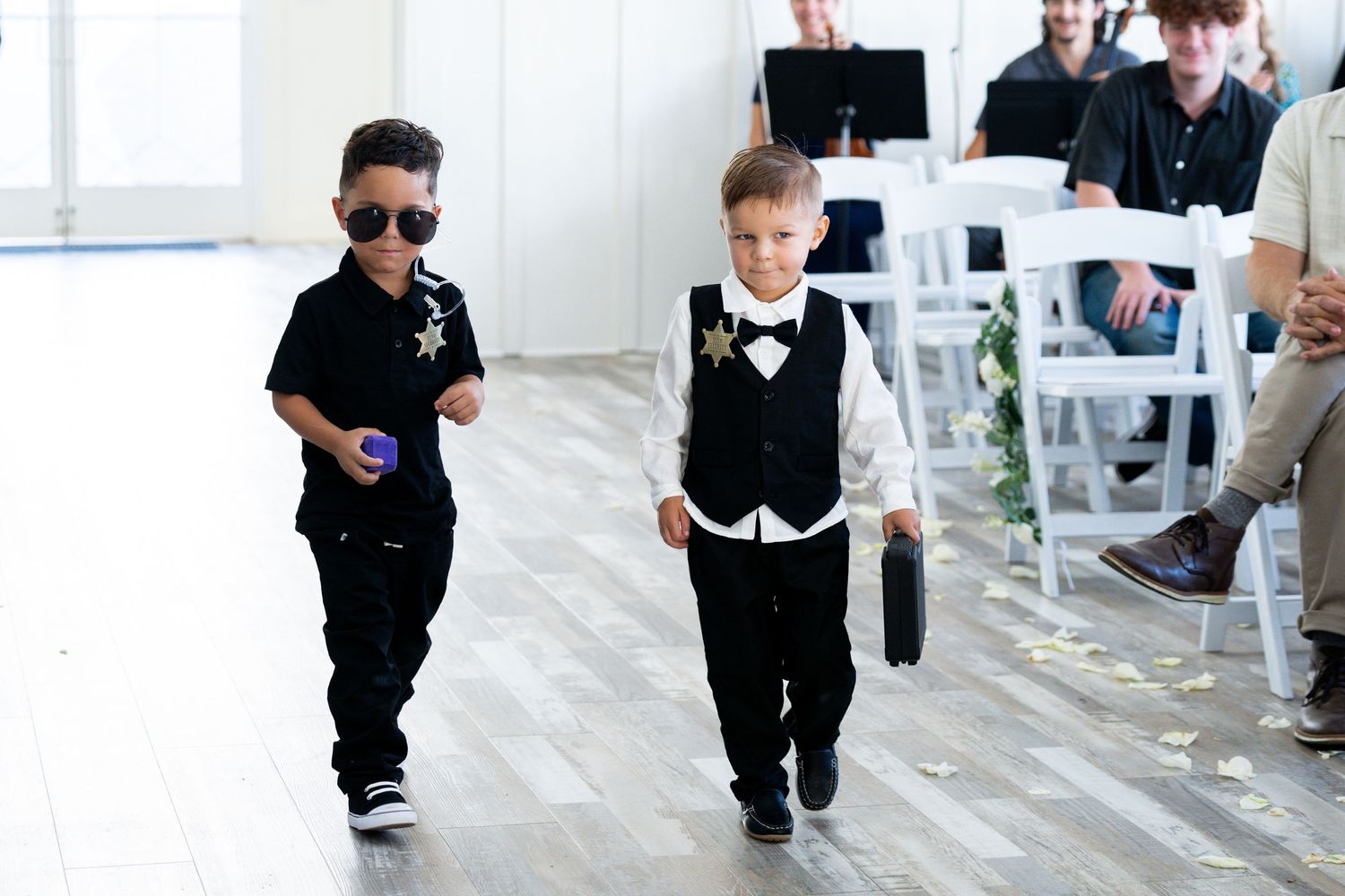 Two young boys in formal black and white outfits walking together at an event with guests seated in the background.