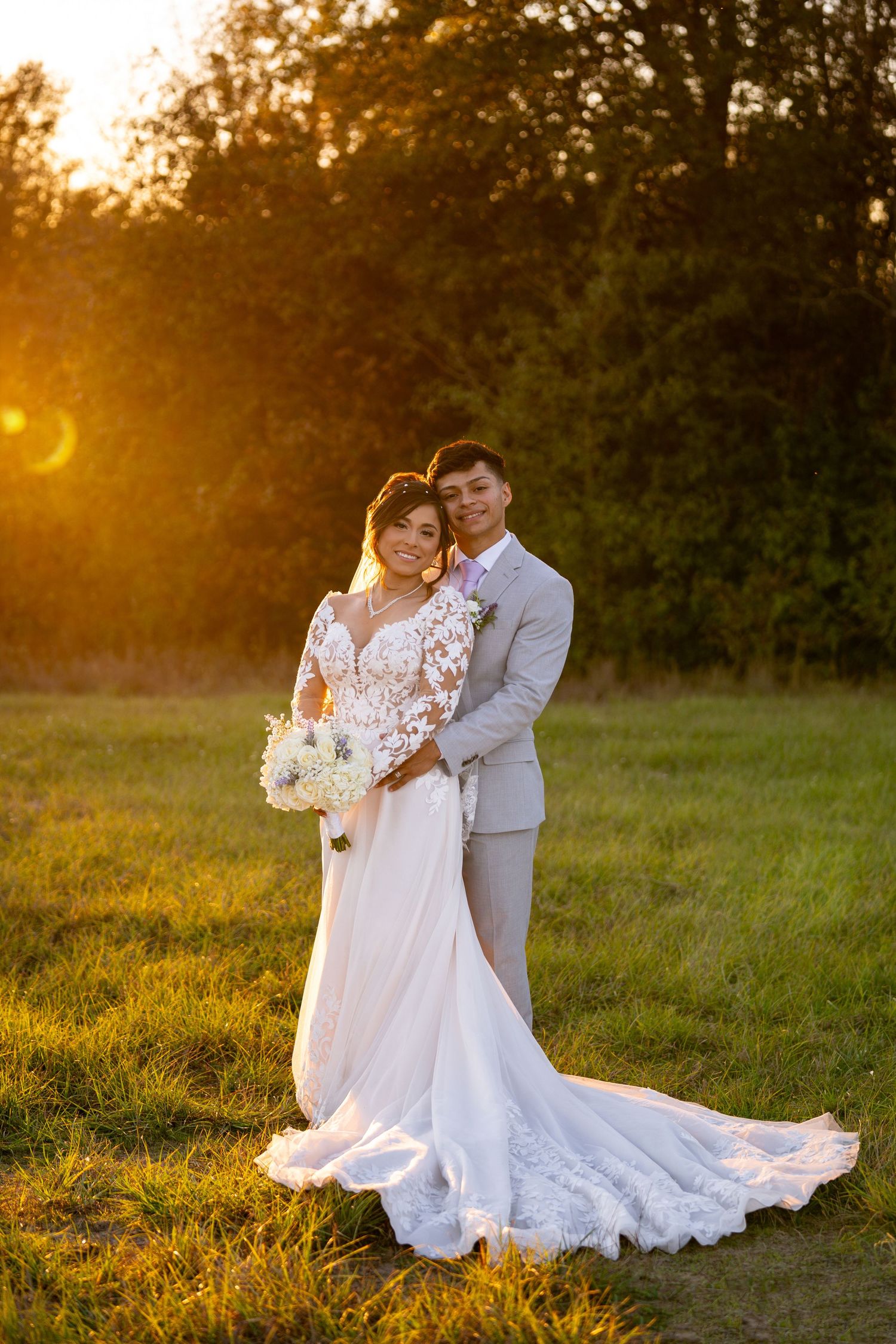 Bride in flowing white lace gown and groom in light gray suit embrace during sunset portraits.