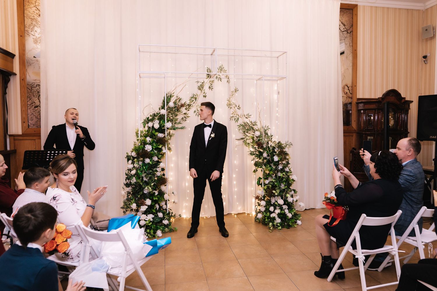 A wedding ceremony in a bright white room with floral arrangements and guests seated in white chairs.
