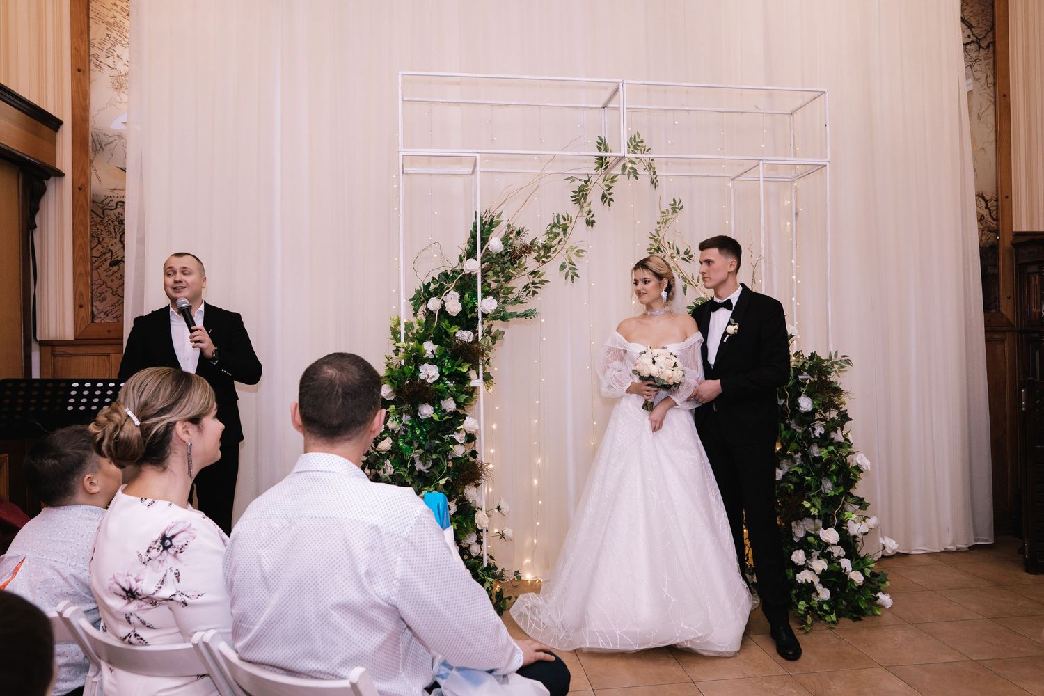 A wedding ceremony takes place in front of a white decorative arch adorned with greenery and florals.
