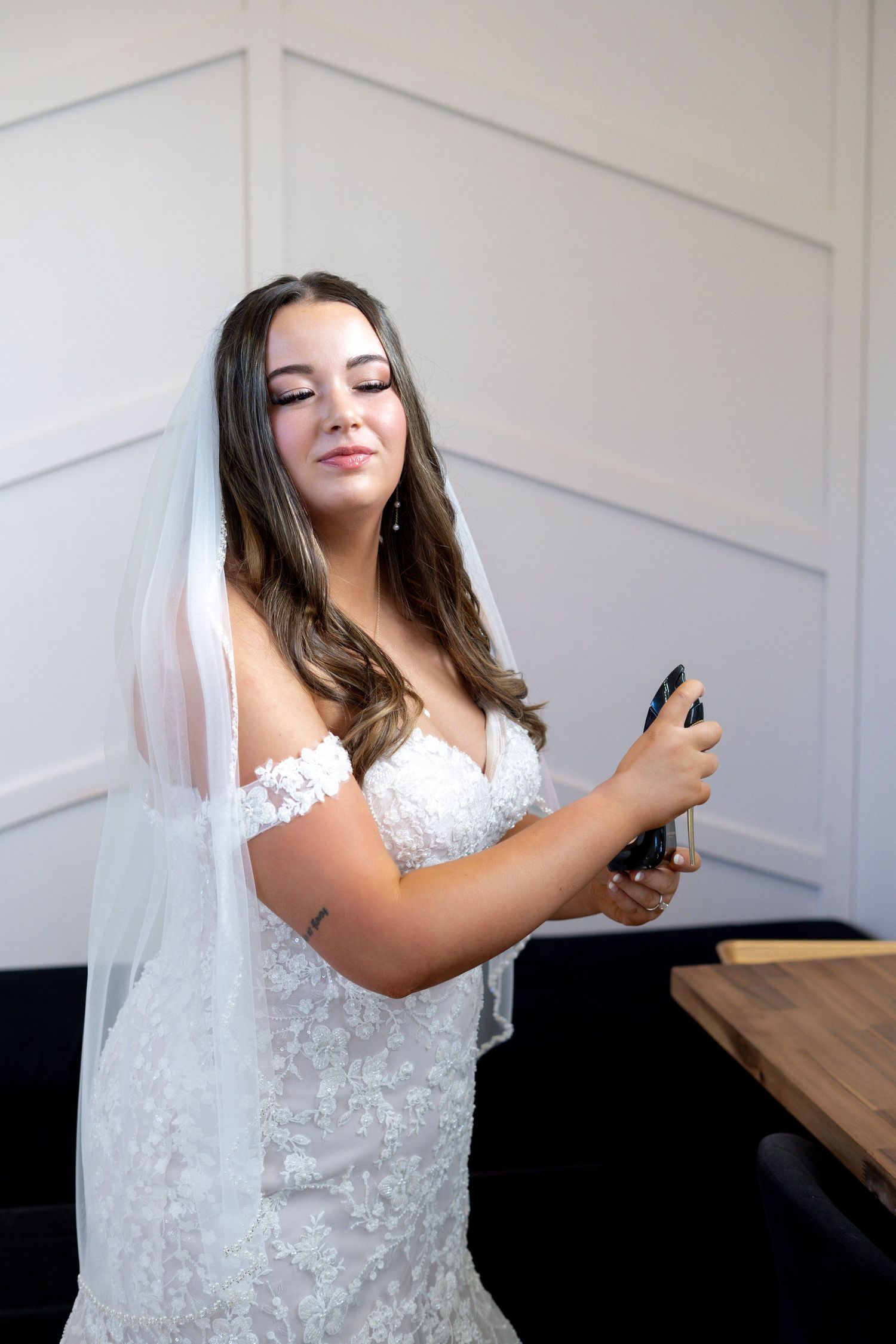 A bride in a white lace wedding dress and veil holds a champagne bottle while posing against a light colored wall.