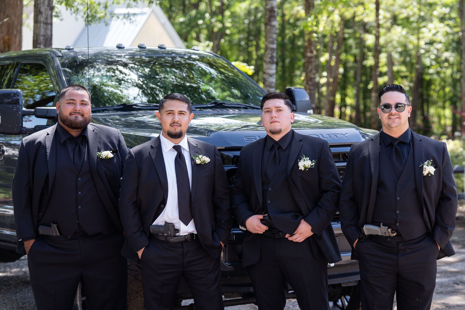 Group wearing black formal attire poses outdoors near parked vehicles in wooded setting.