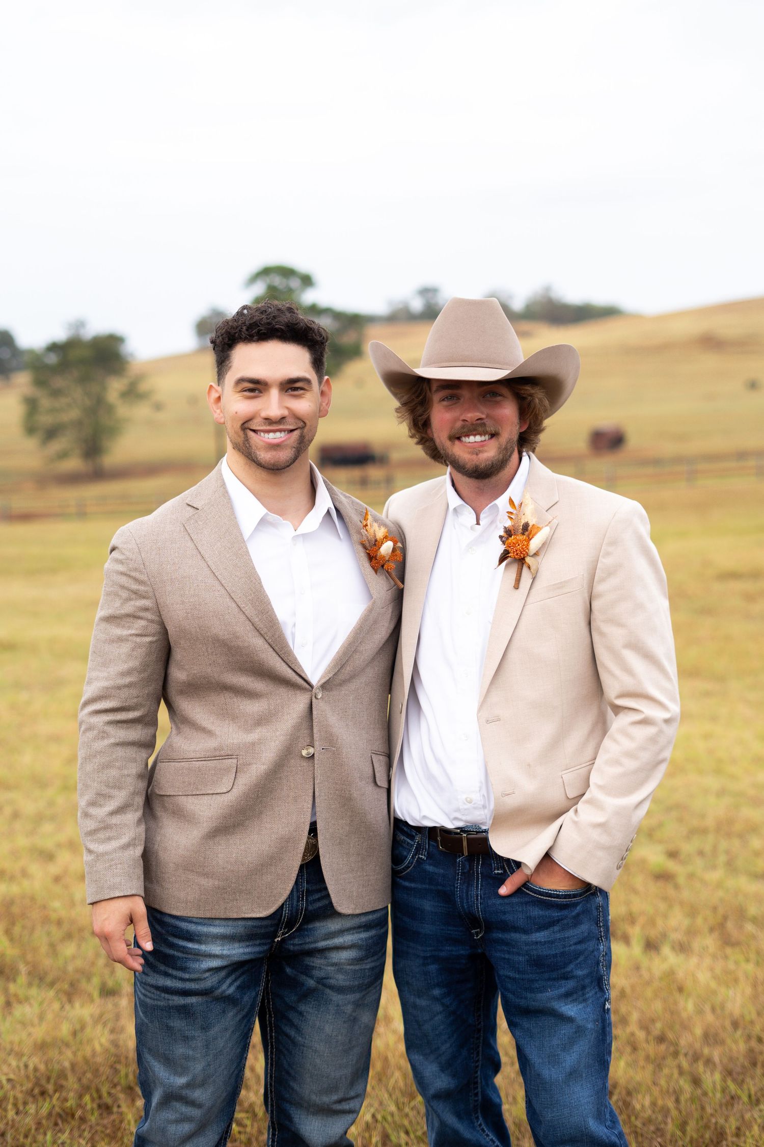 Two men in light colored suits and boutonnieres pose together in a rustic field setting.