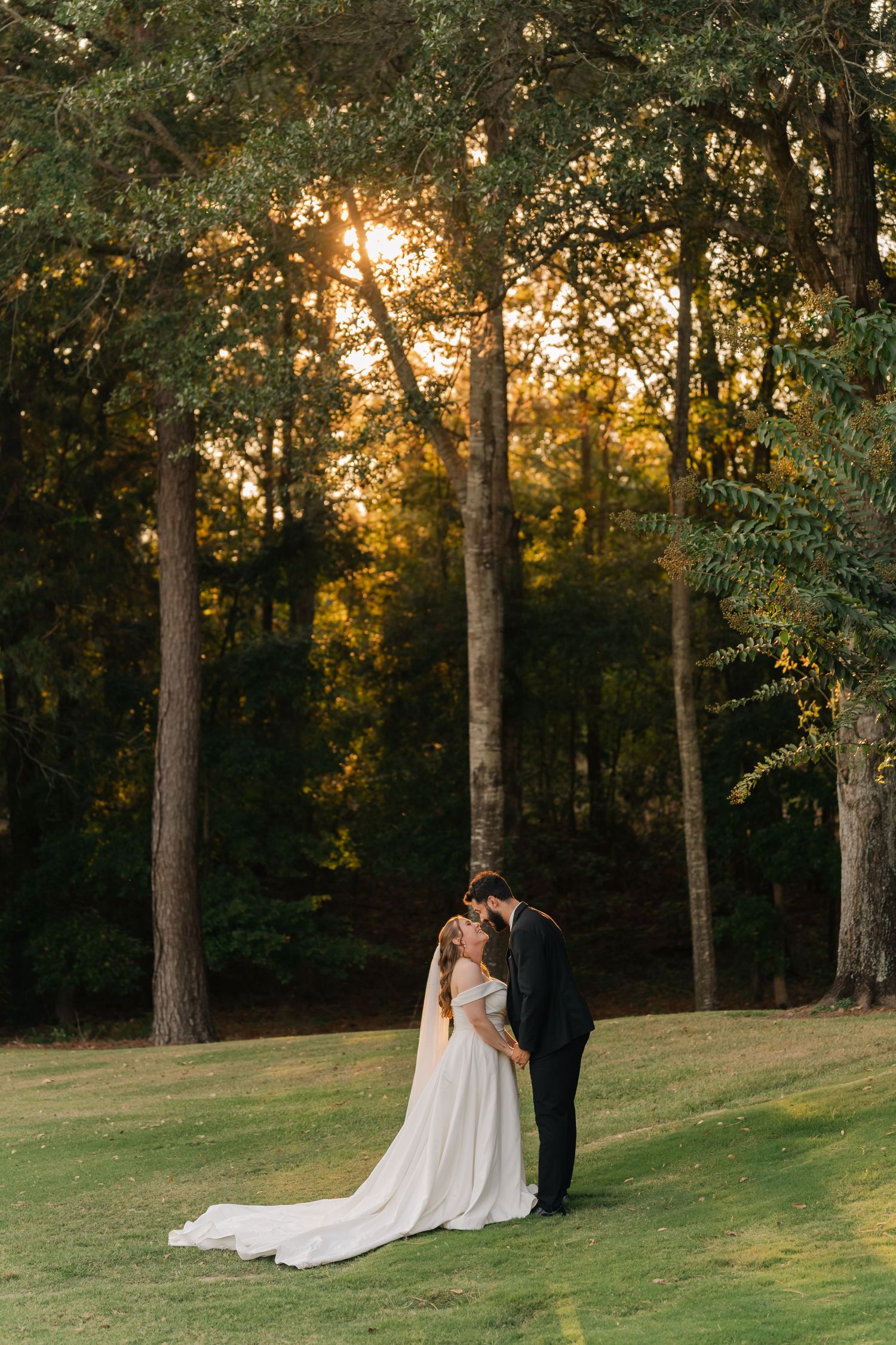 A couple shares an intimate moment at sunset on a grassy hill surrounded by tall trees.
