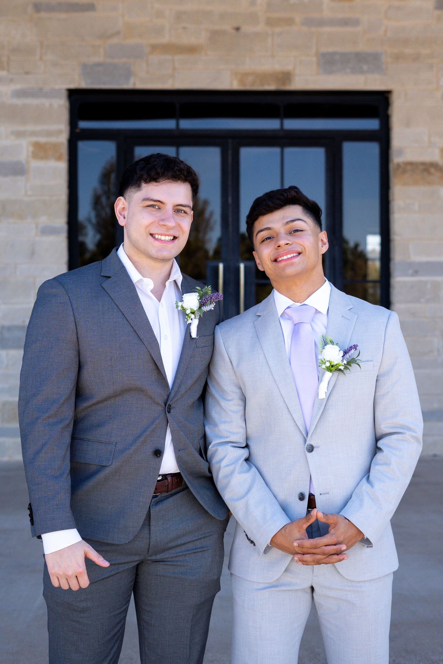 Wedding party members in coordinated grey suits with white boutonnieres pose against brick wall.