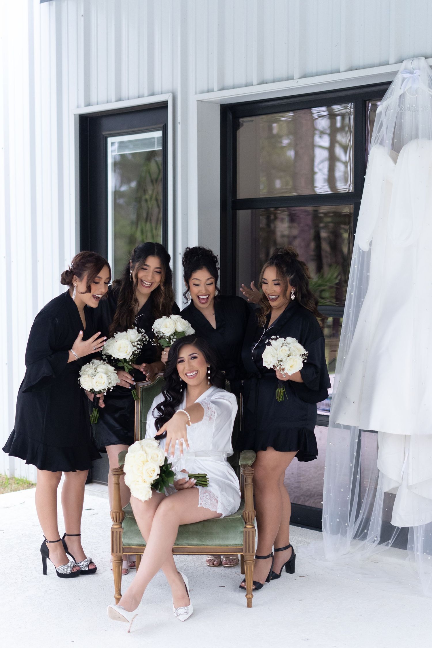 Wedding party in black dresses and white bouquets pose playfully for photos in a bridal suite.