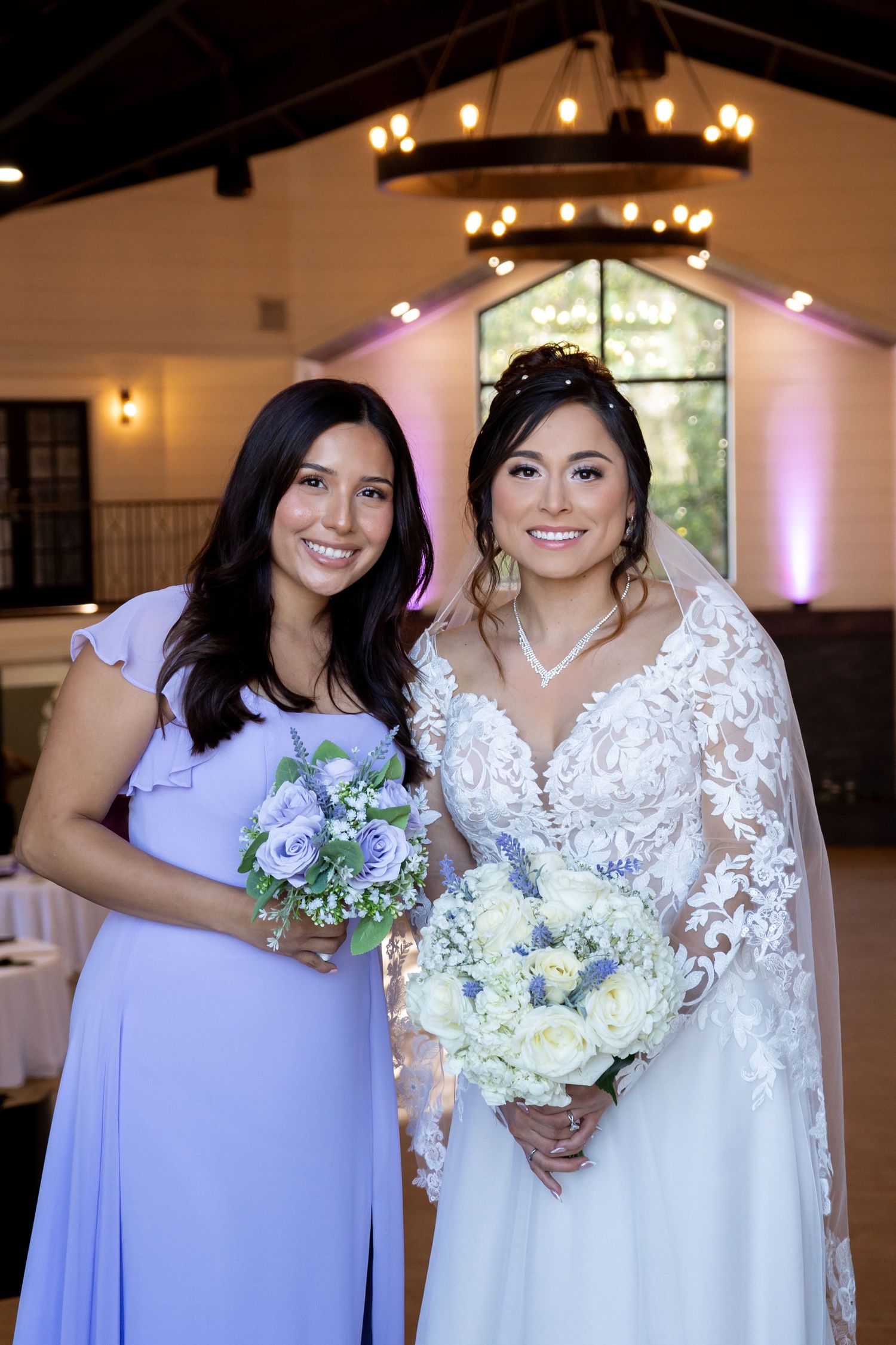 Bridesmaids in light purple dresses pose with bride wearing lace wedding gown at indoor venue with chandelier lighting.