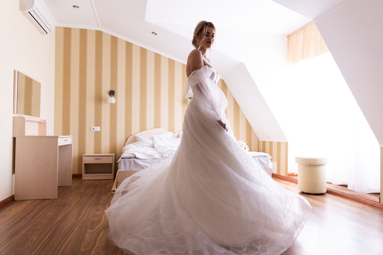 A bride in a flowing white wedding gown poses gracefully in a bright interior hallway with striped wallpaper.