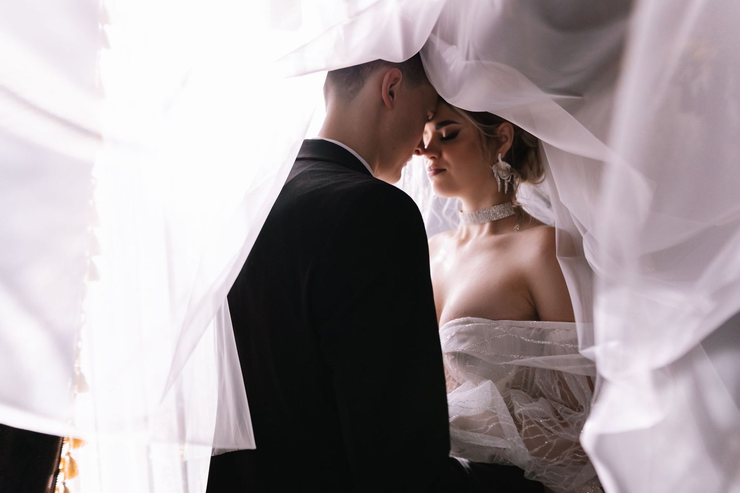 A romantic wedding couple embraces under flowing white veils during an intimate photoshoot moment.
