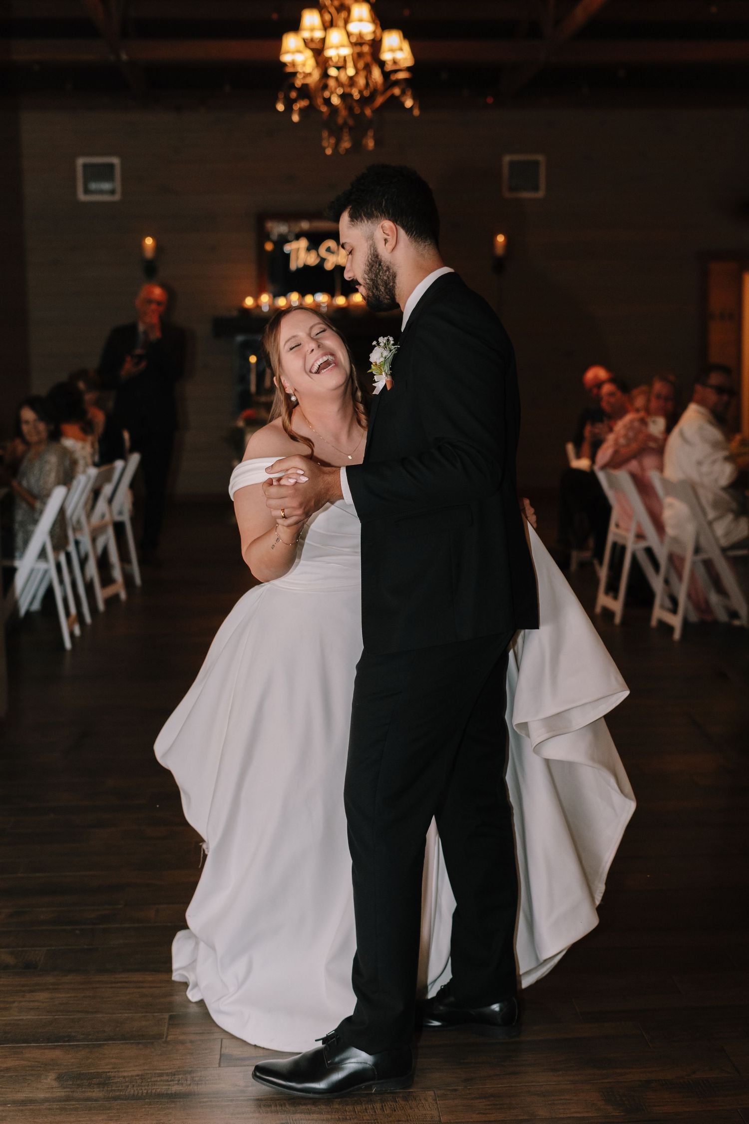 A couple shares their first dance on a dark wooden floor under warm chandelier lighting.