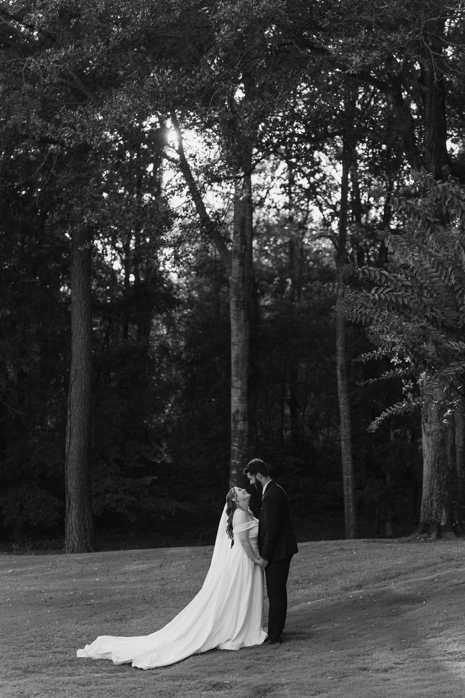 A romantic wedding couple poses together on a grassy hill surrounded by tall trees in a black and white photo.