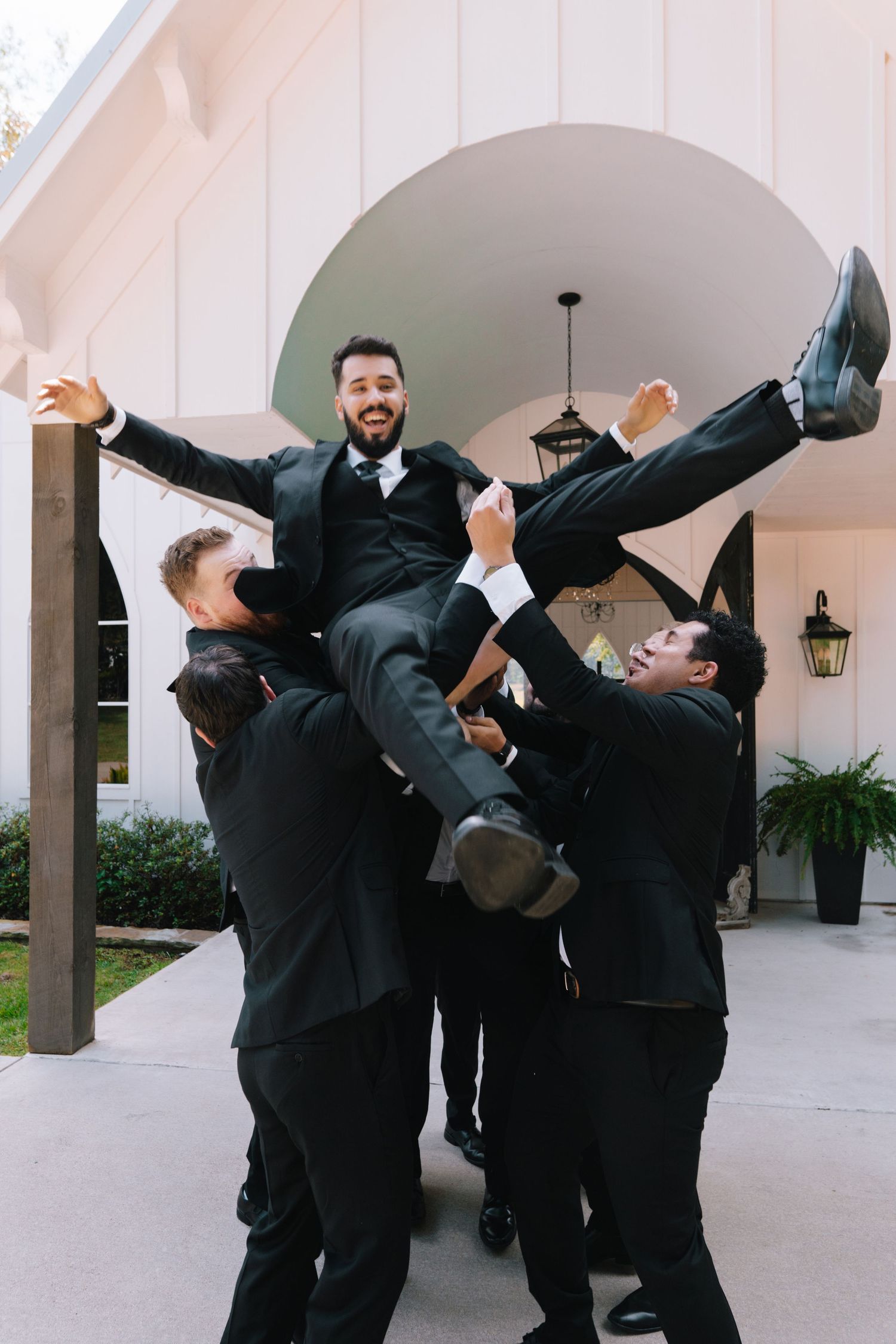 Groomsmen in black tuxedos strike playful poses and dance moves under an outdoor archway.