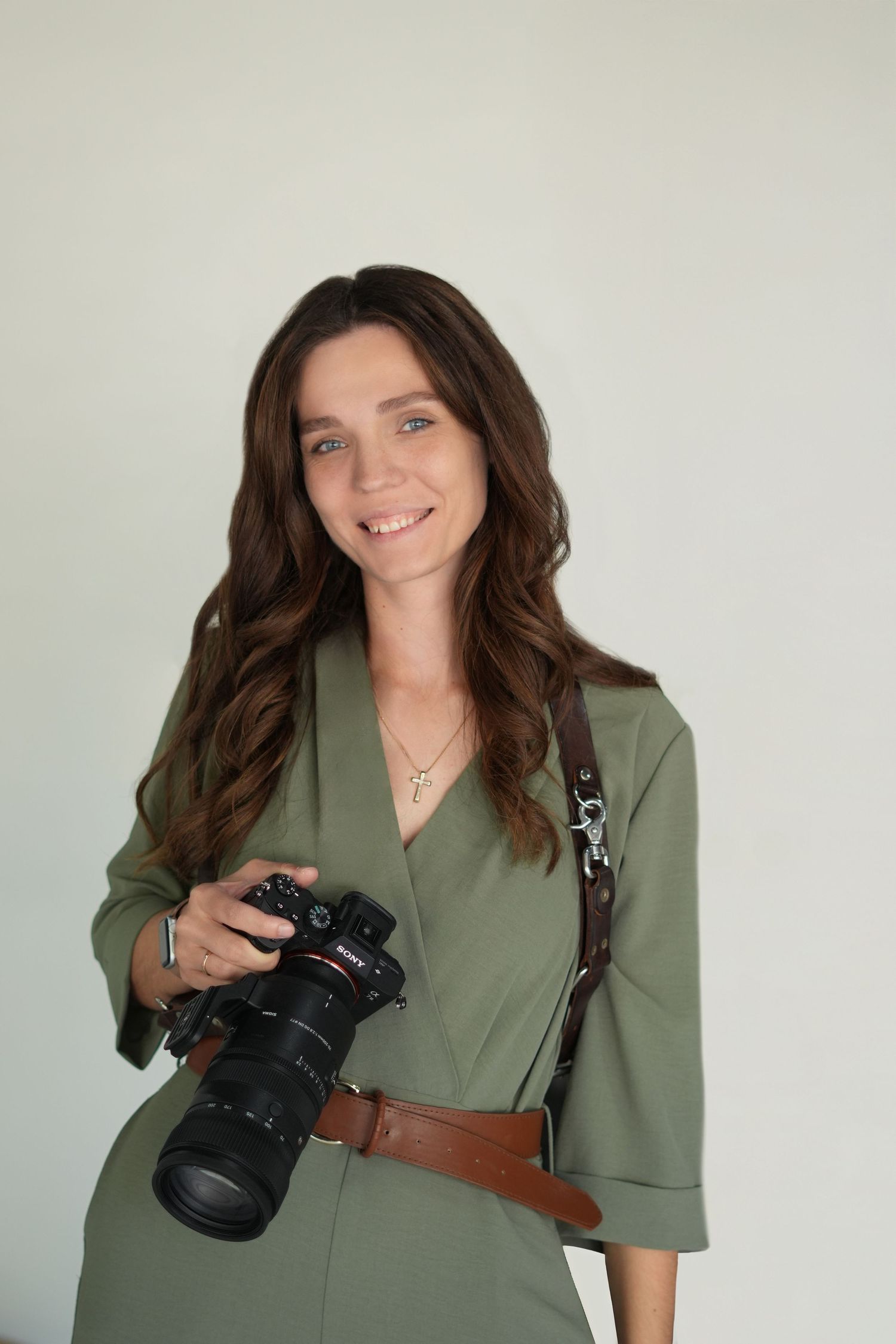 A smiling woman with long brown hair holds a DSLR camera with a leather strap, wearing a green wrap dress.