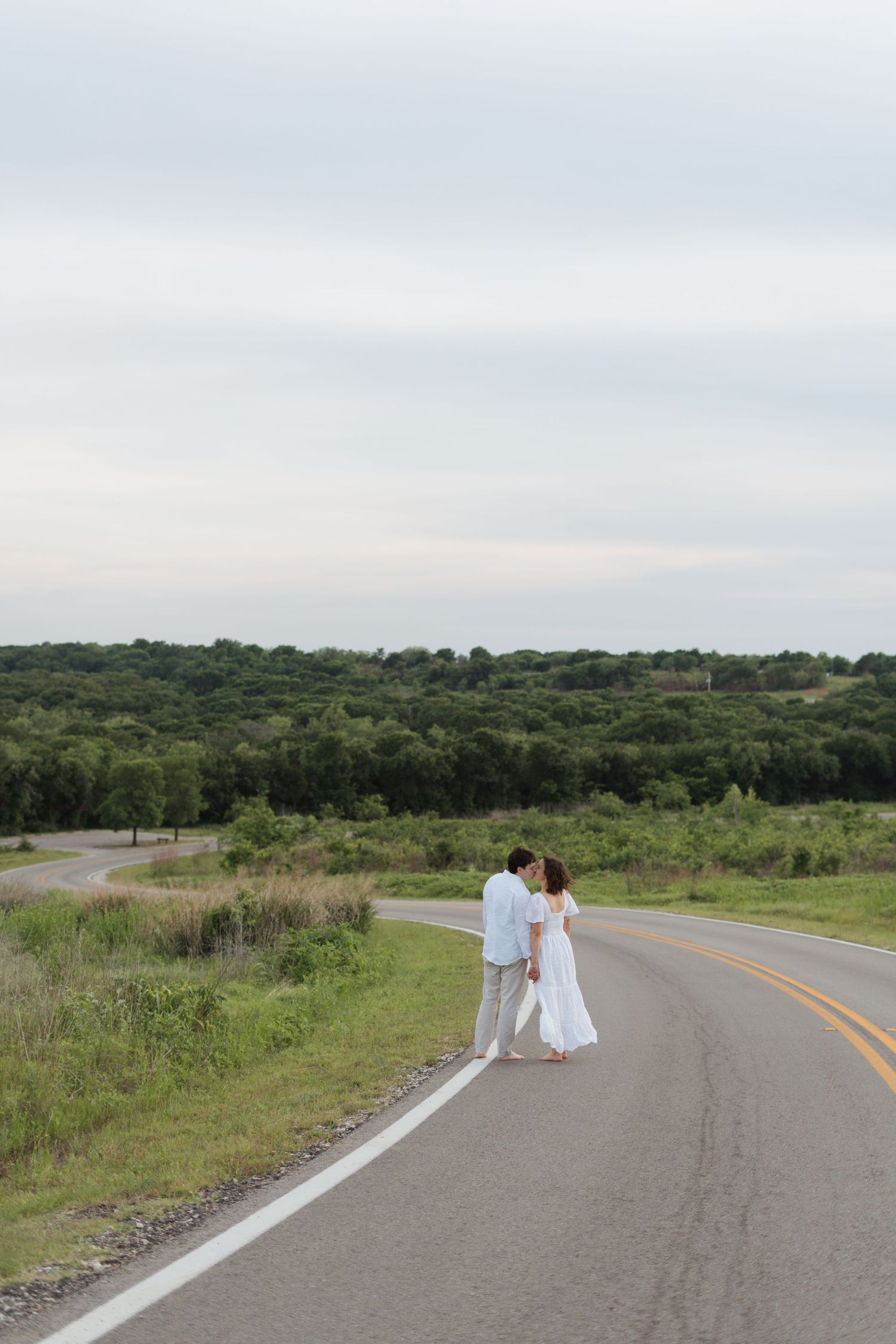 Couple in white outfits standing on a rural road surrounded by green trees and fields under a cloudy sky.