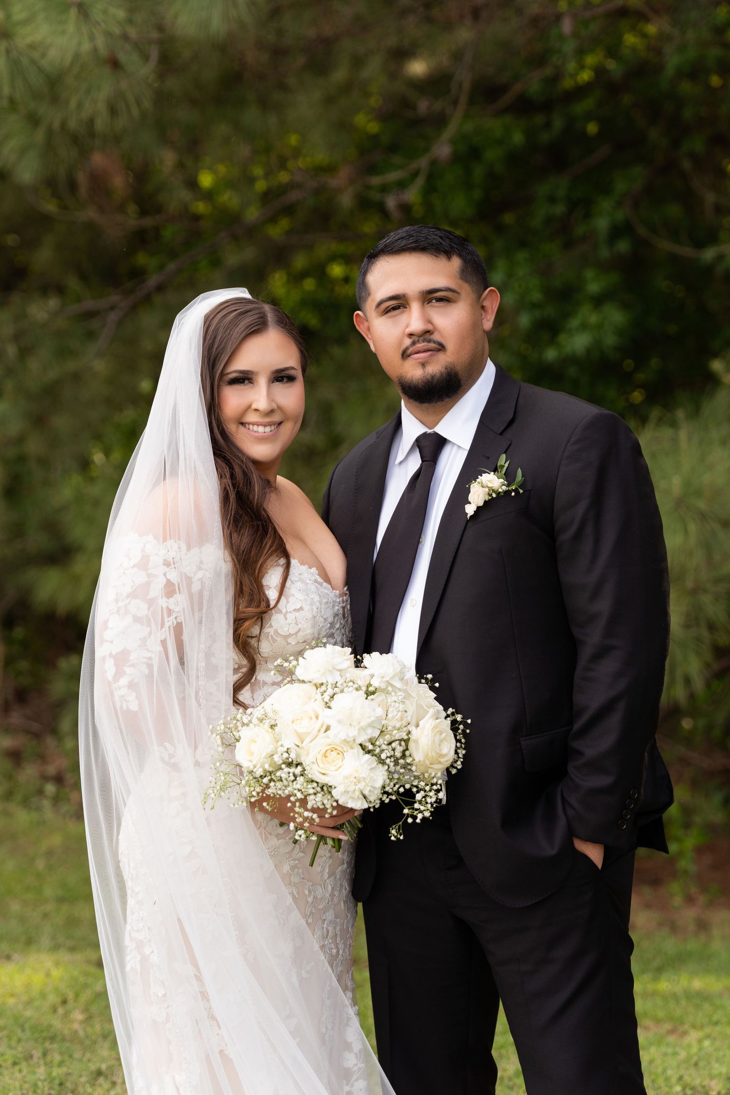A bride and groom pose together outdoors in formal wedding attire with white floral bouquet and natural backdrop.