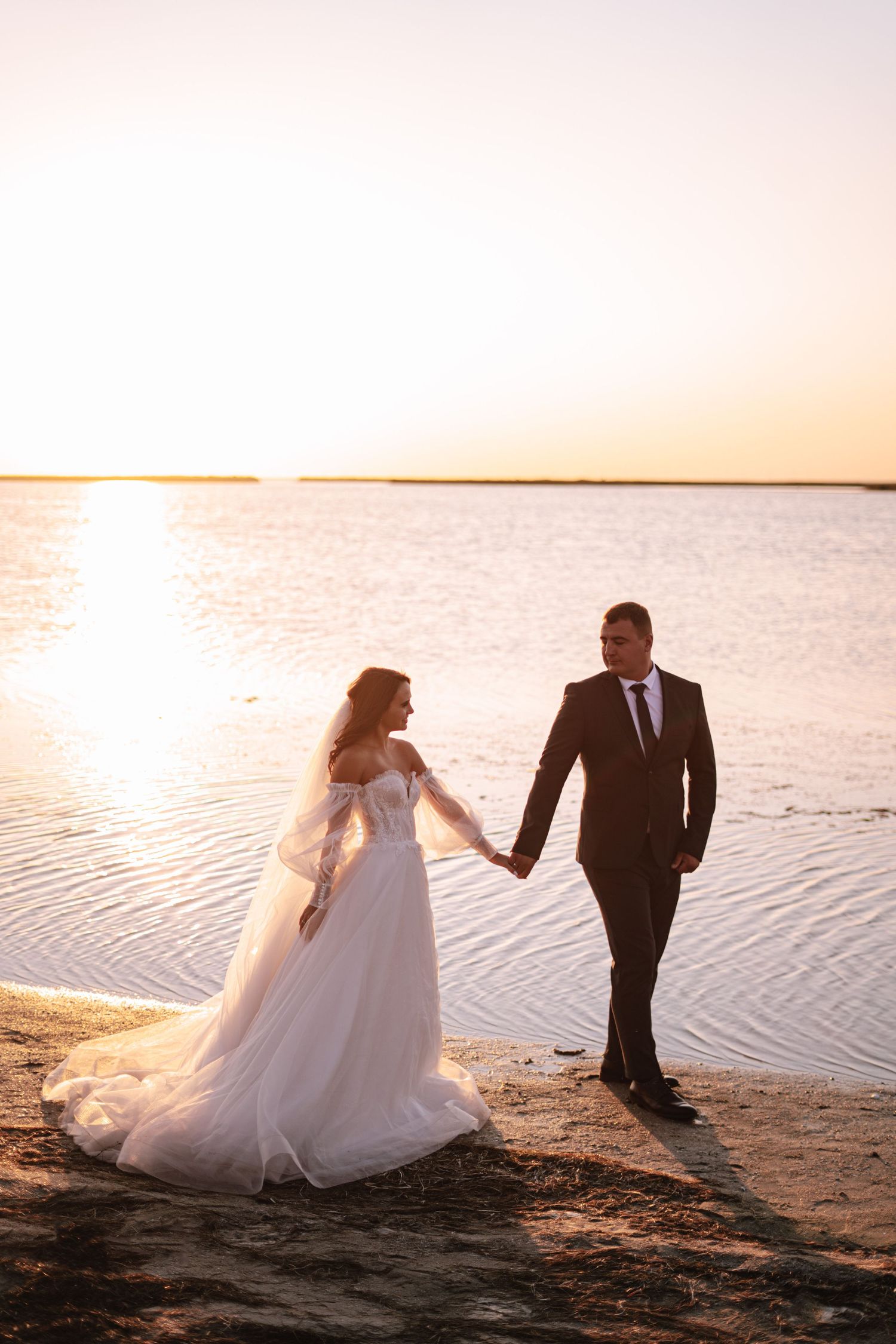 A couple in wedding attire holds hands while walking along a beach shoreline at sunset.