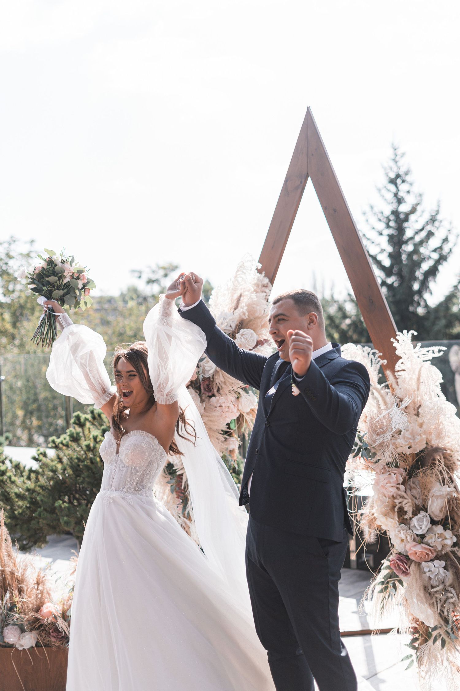 A bride and groom celebrate joyfully under an outdoor wedding arch decorated with white and blush floral arrangements.