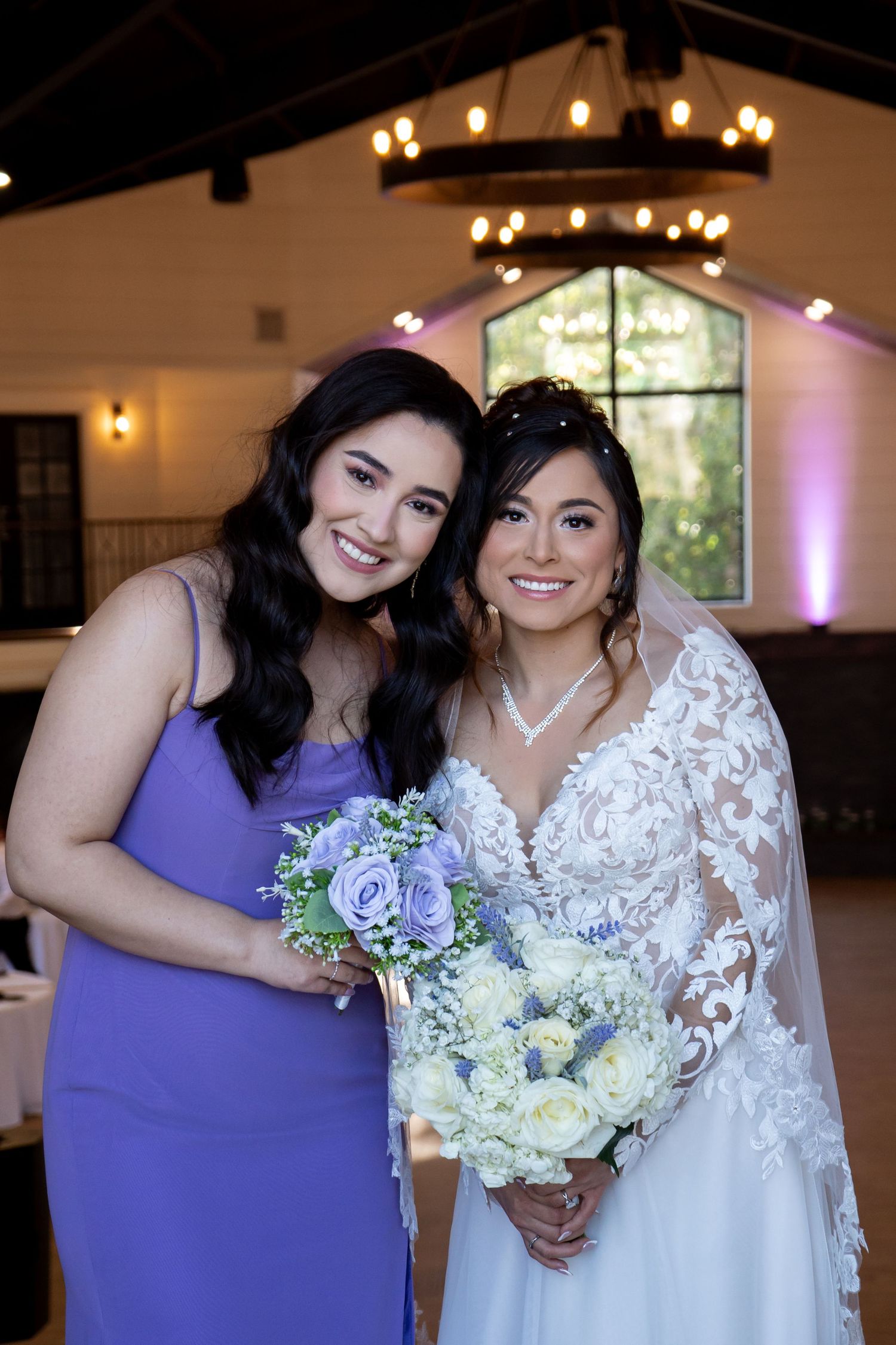 Wedding party members pose together holding purple and white floral bouquets in an elegant venue.