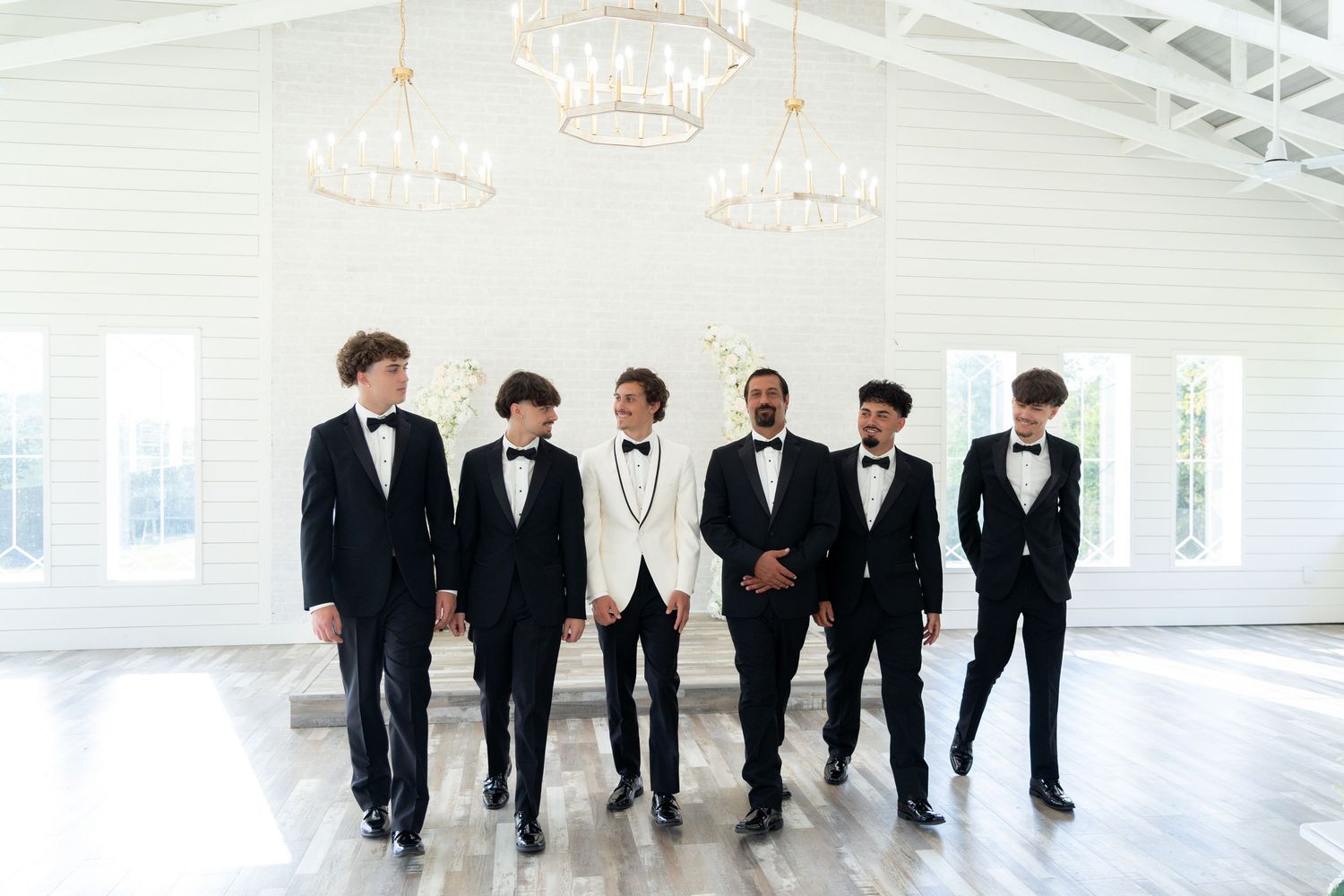 A group of six people in formal black and white attire walk together in an elegant white room with chandeliers.