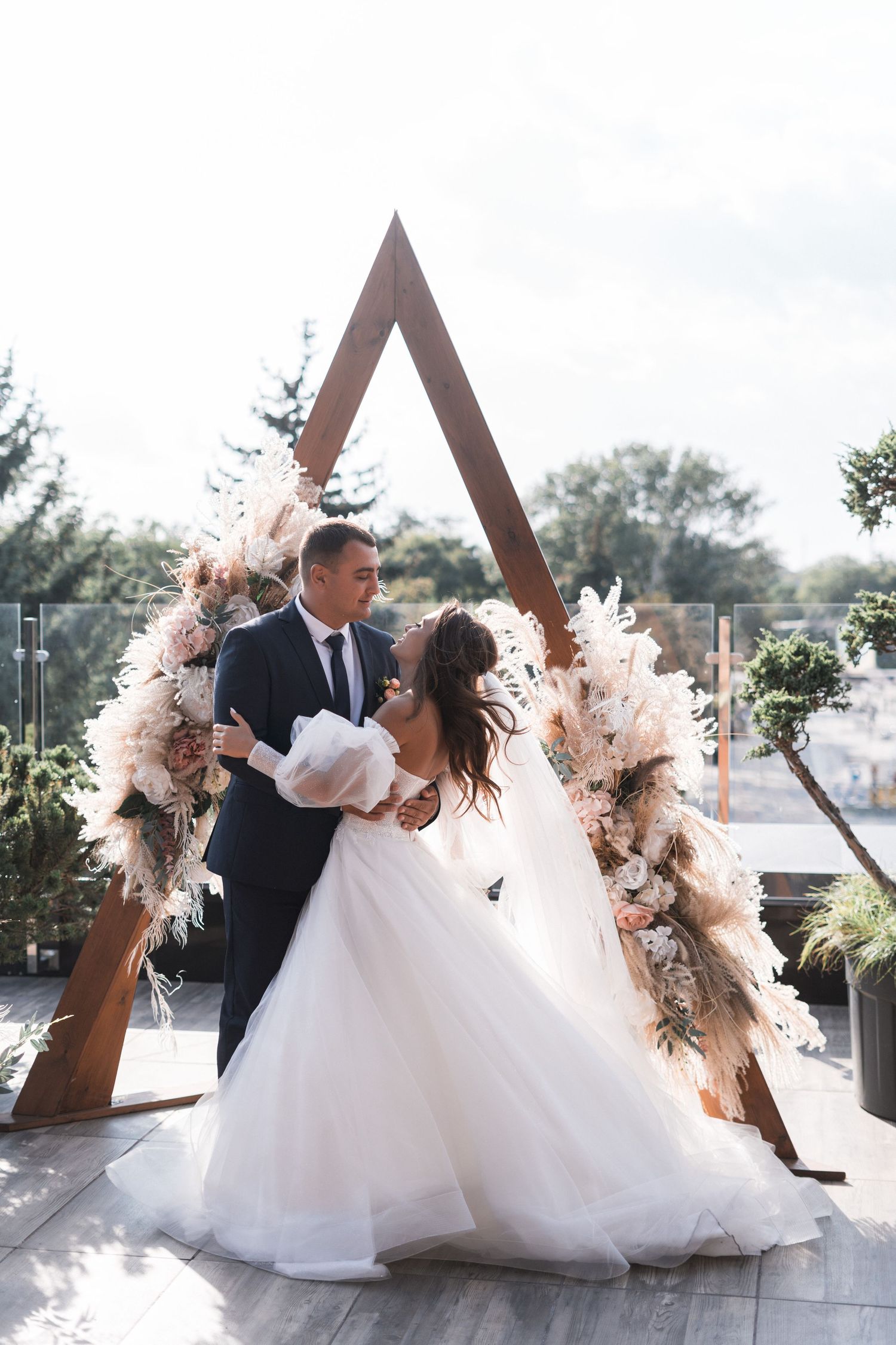 Couple embraces under a triangular wooden arch decorated with blush pink pampas grass and dried floral arrangements.