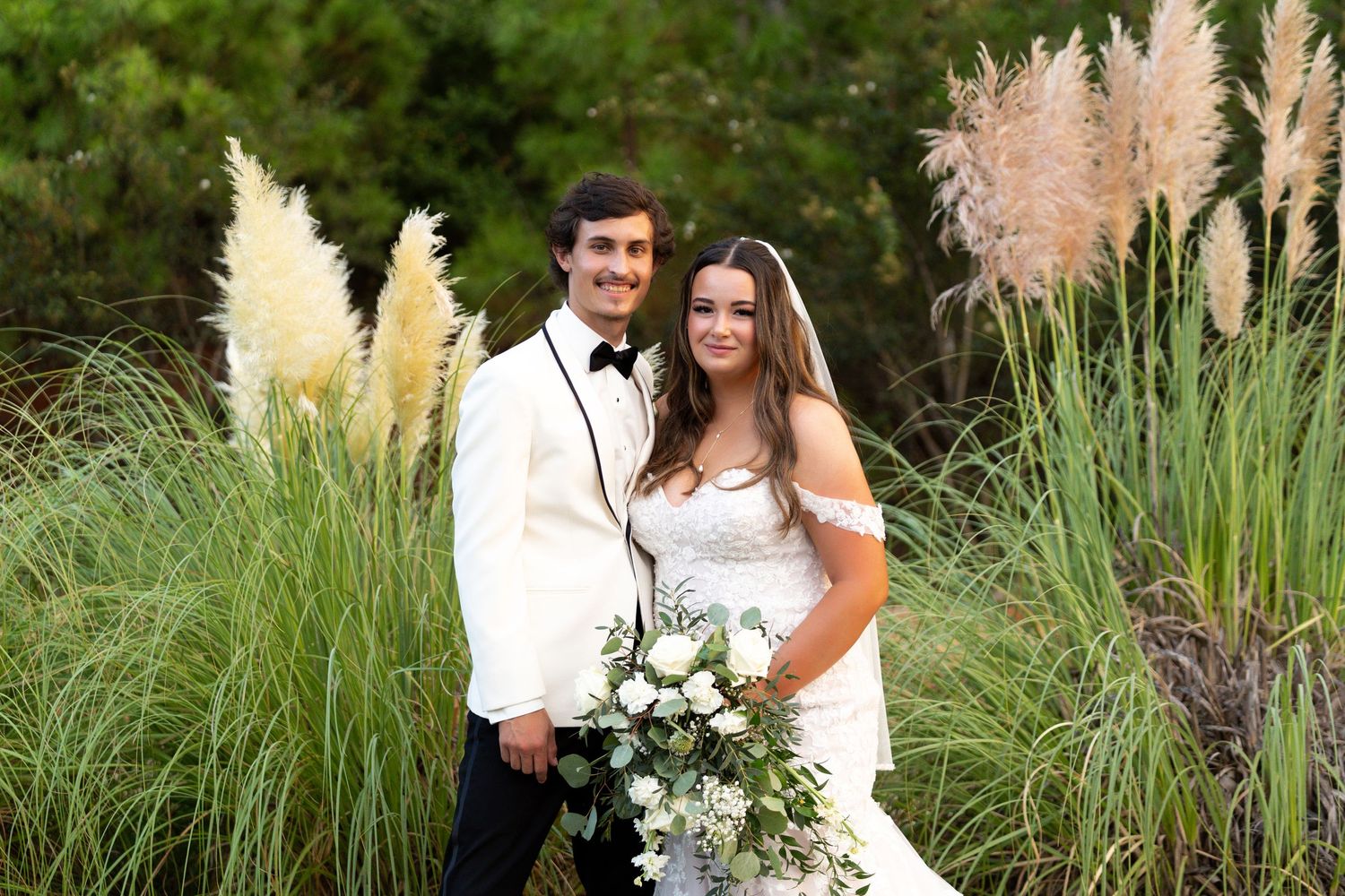 A couple poses together in their wedding attire in front of tall ornamental pampas grass on their special day.