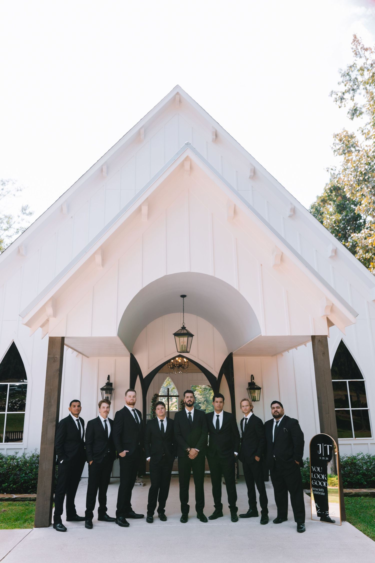 Groomsmen in black suits line up in front of a white chapel with gothic arched windows and lanterns.