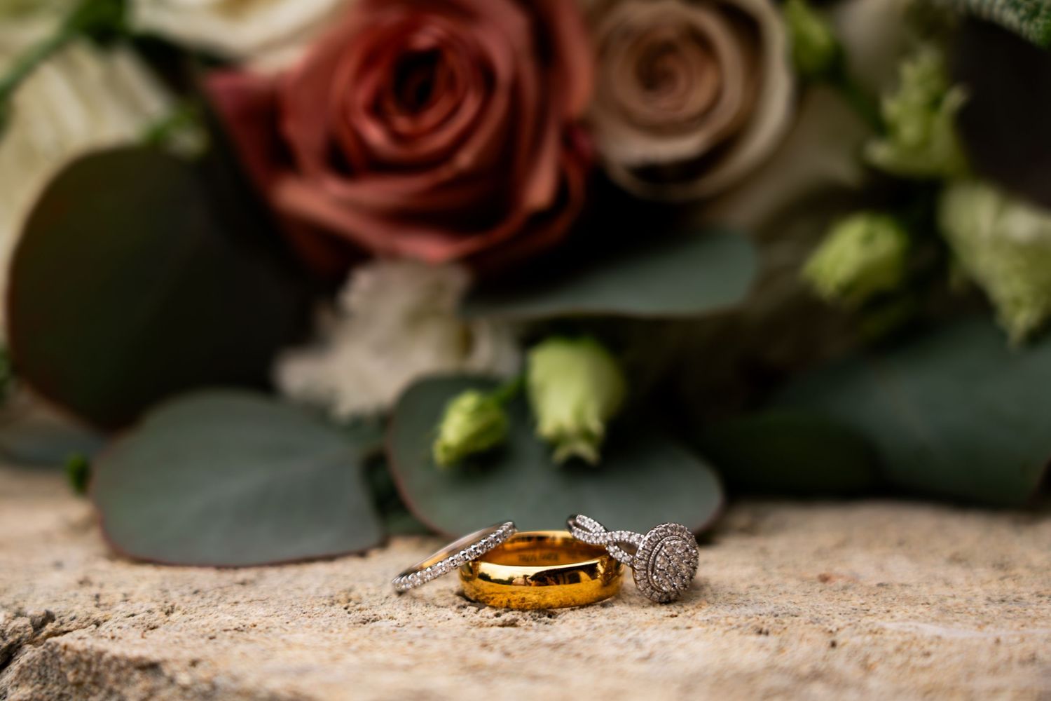 Close-up detail shot of pink roses and wedding rings on textured stone surface.
